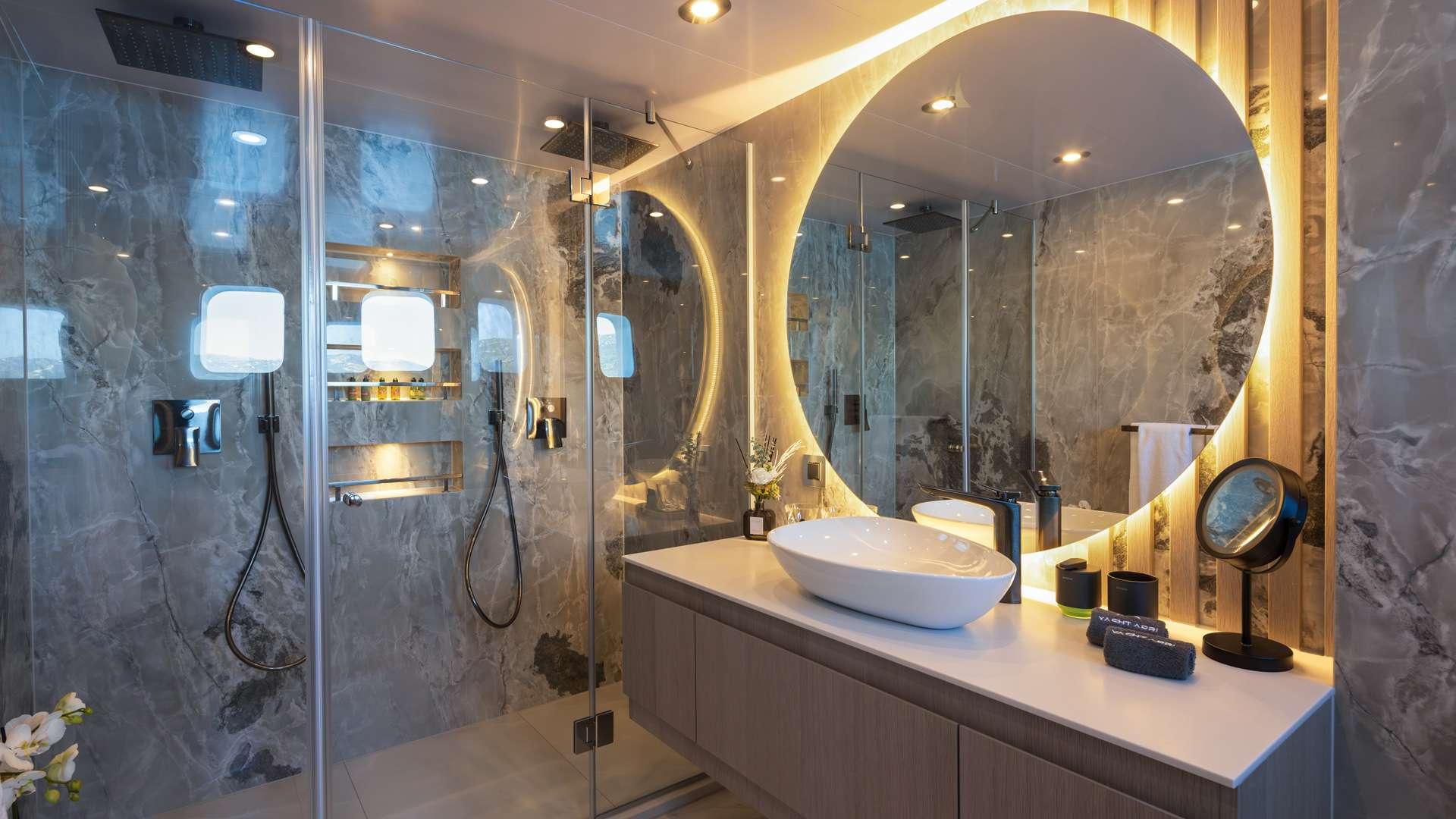 luxury yacht en-suite bathroom featuring marble walls, white vessel sink, backlit mirror and glass shower