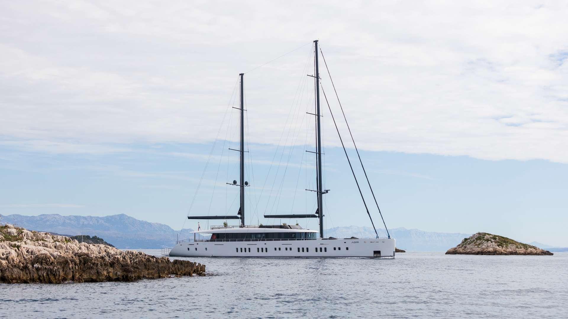 Large white sailing yacht with two masts anchored between rocky coastline and mountains
