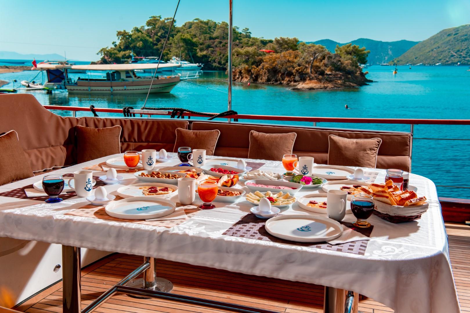 yacht aft deck dining table with breakfast spread, cushioned seating, overlooking turquoise bay