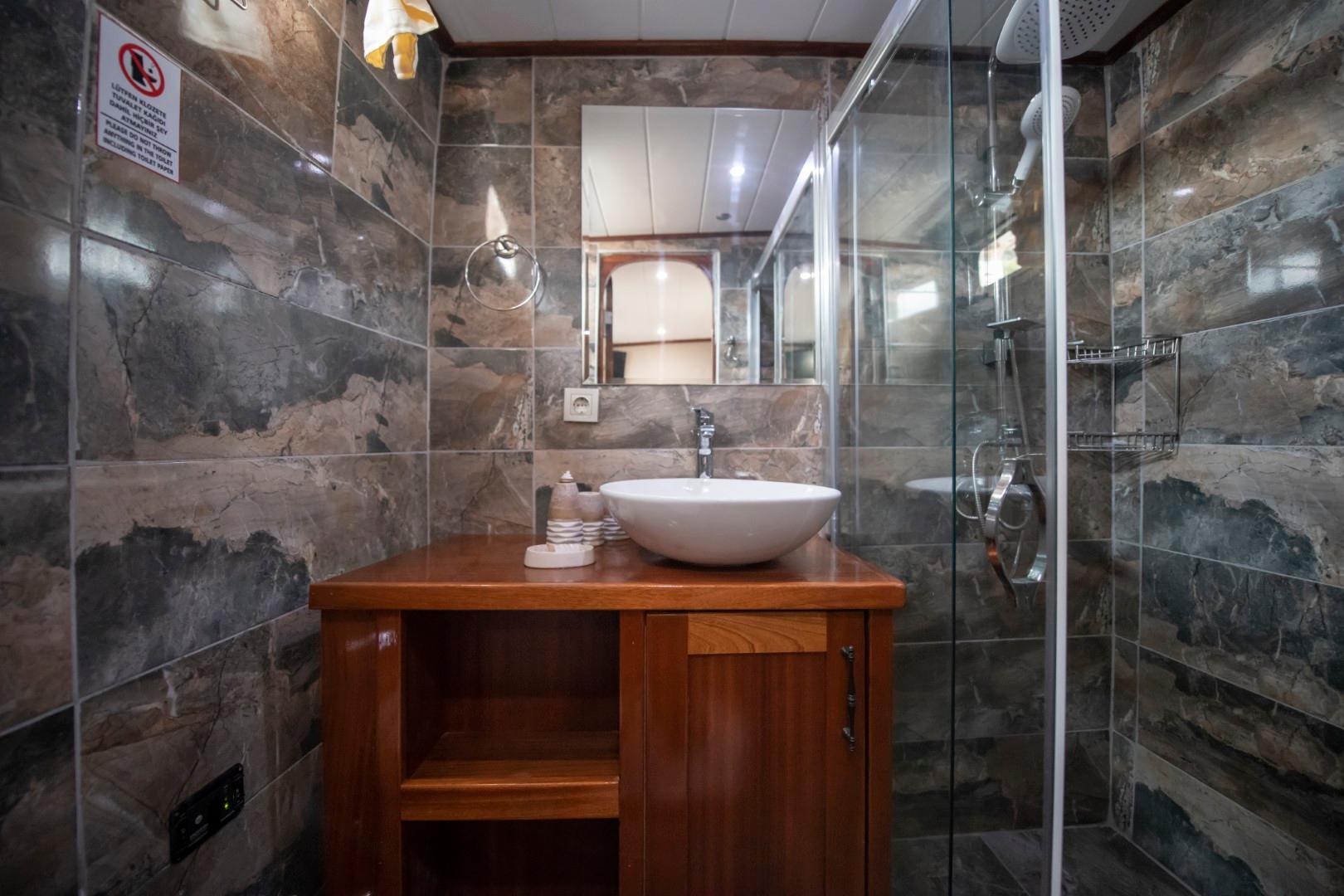 yacht bathroom with dark marble tile walls, white vessel sink on wooden vanity, glass shower enclosure