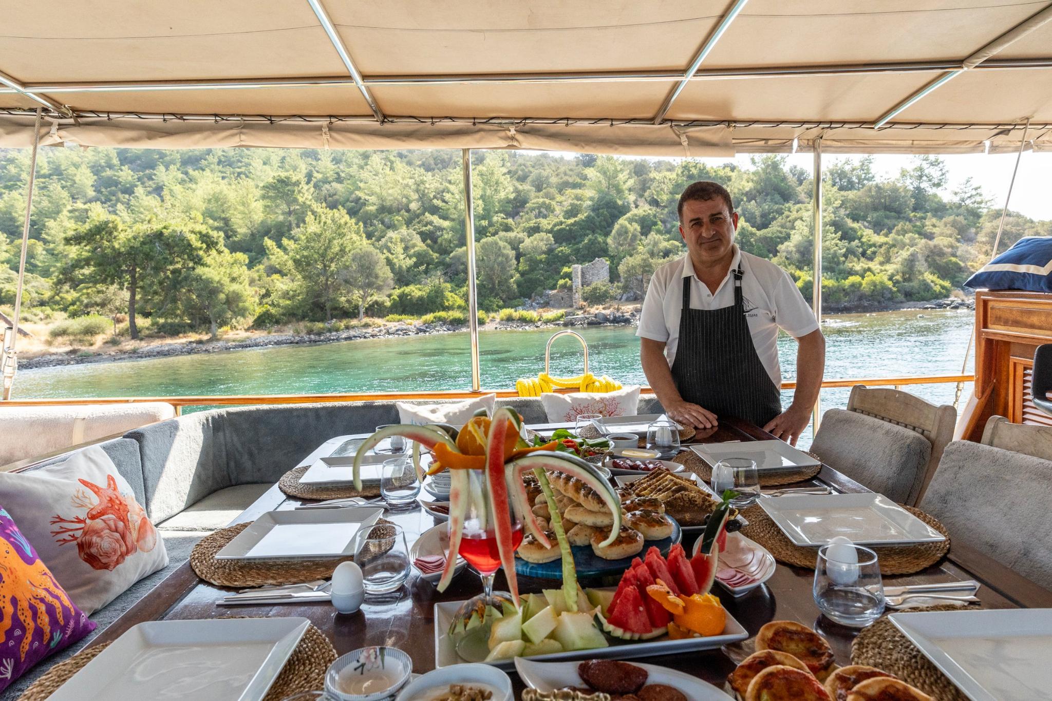 yacht chef in apron presenting breakfast spread with fresh fruit and pastries on covered aft deck