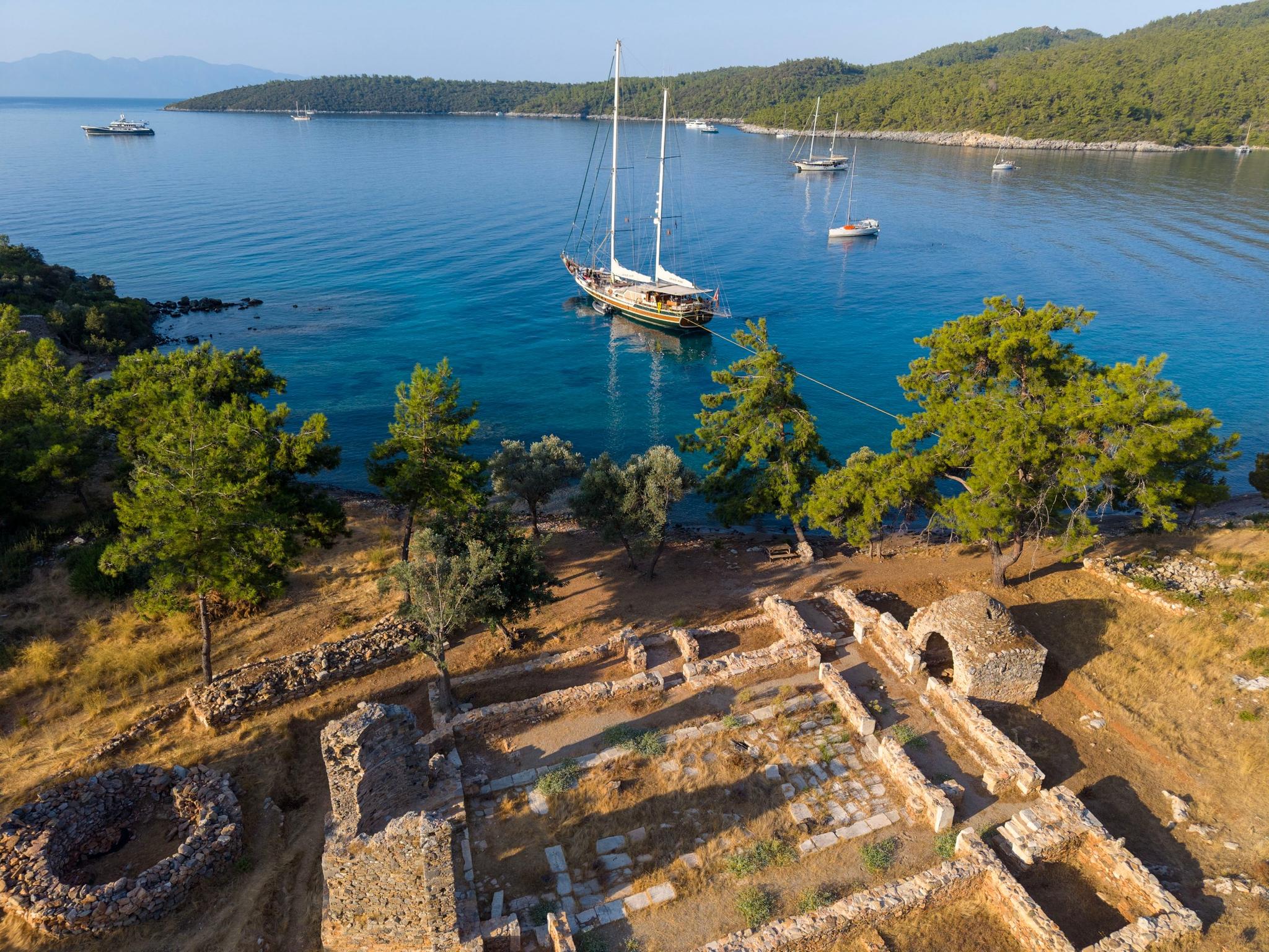 Aerial view of traditional wooden gulet anchored in turquoise bay near ancient stone ruins and forested coastline