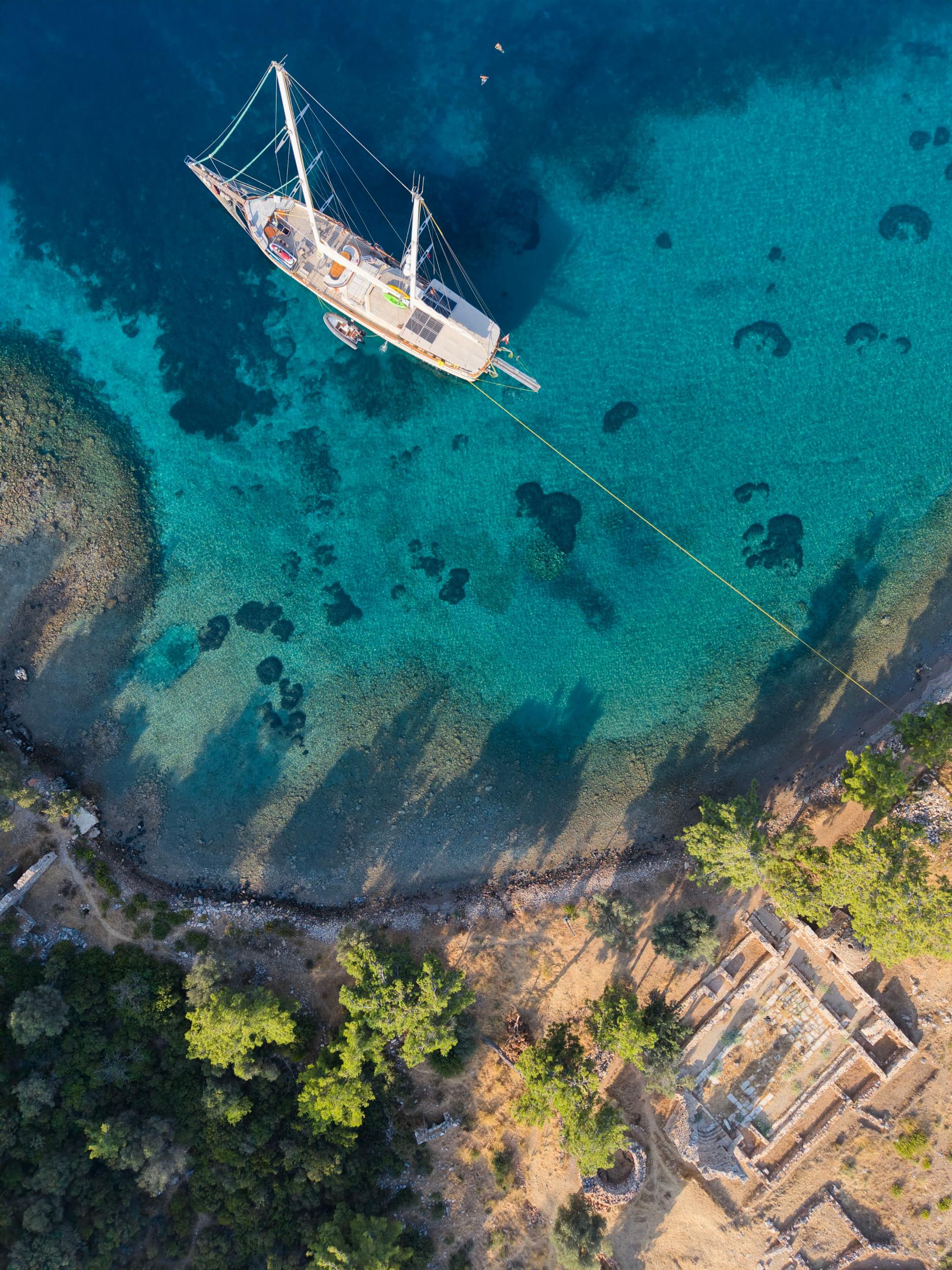 aerial view of white gulet yacht anchored in crystal clear turquoise bay near ancient stone ruins