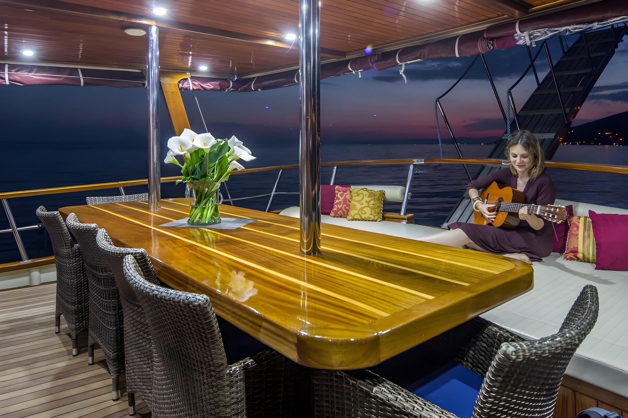 Aft deck outdoor dining table with wicker chairs and guest playing guitar at evening