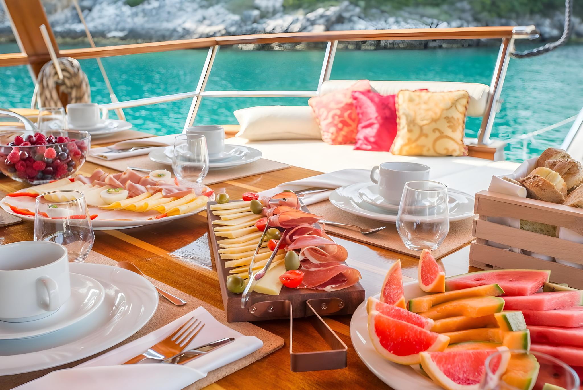 outdoor breakfast table with fresh fruit, charcuterie, bread and table settings on yacht deck