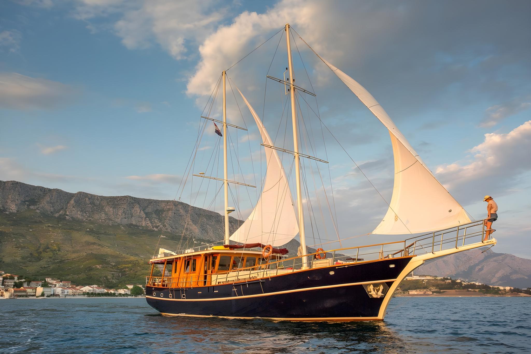 traditional wooden gulet with white sails and crew member on deck sailing near mountainous coastline
