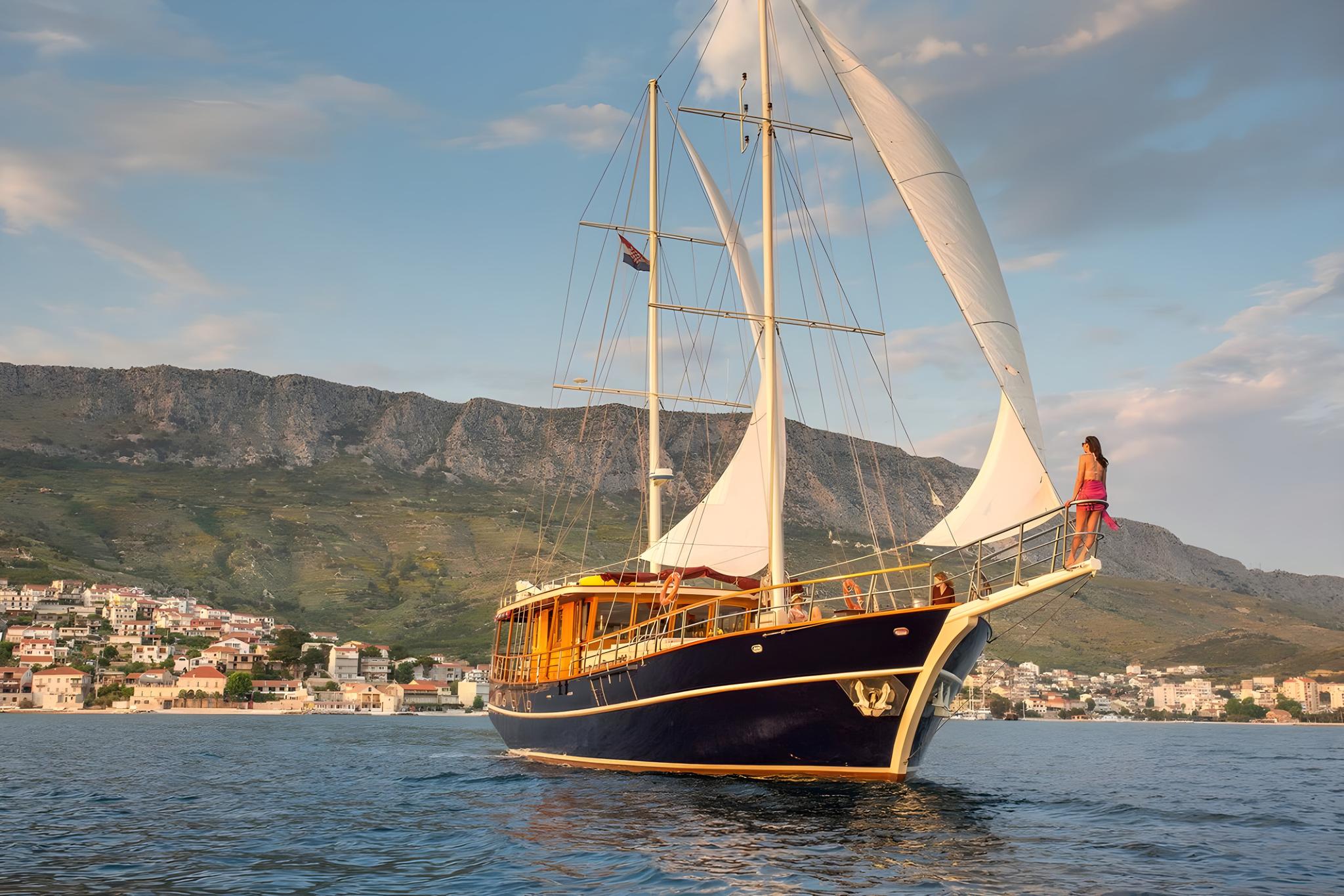 Classic wooden gulet with dark blue hull and cream sails cruising near Mediterranean coastline