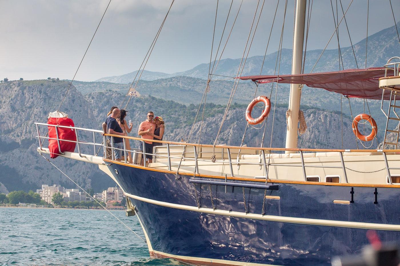 wooden gulet yacht with guests on deck against mountainous Mediterranean coastline