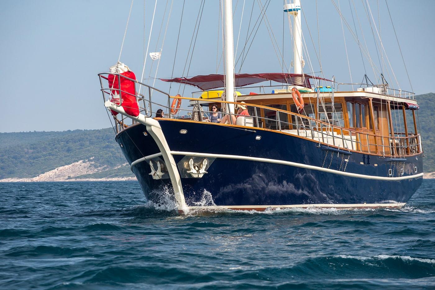 Traditional wooden gulet yacht with dark blue hull and varnished wood superstructure sailing in Mediterranean waters with coastal hills in background