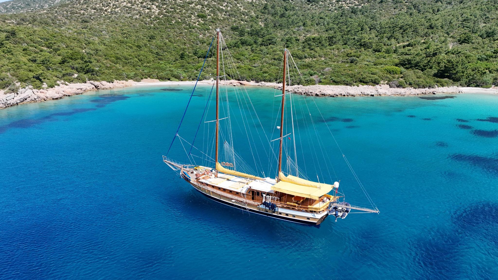 aerial view of traditional wooden gulet with two masts anchored in crystal clear turquoise Mediterranean waters near forested coastline
