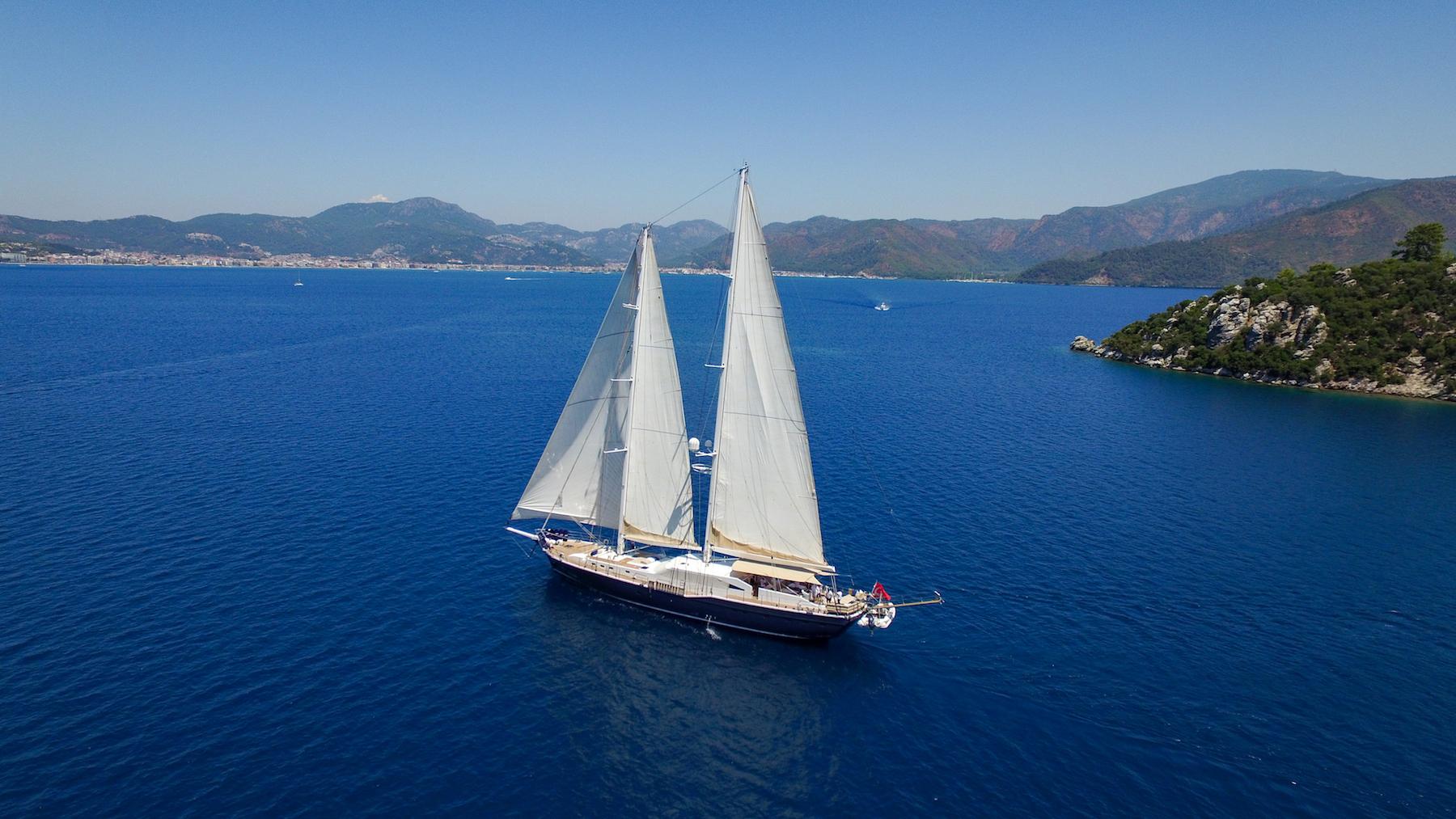 aerial view of luxury sailing gulet with white sails raised cruising blue Mediterranean waters near mountainous coastline