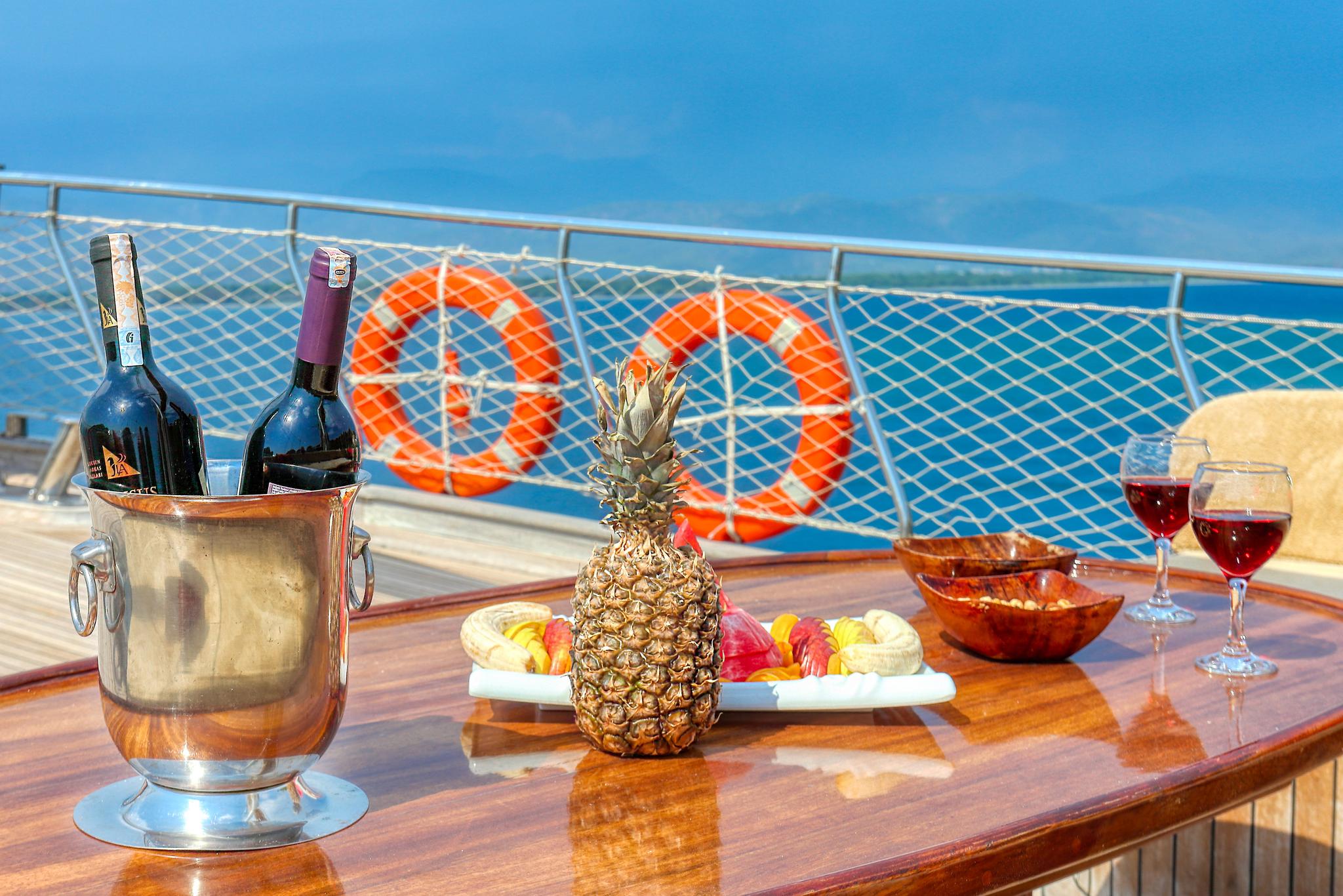 deck dining table with wine bottles, glasses, fresh pineapple and fruit platter aboard luxury yacht