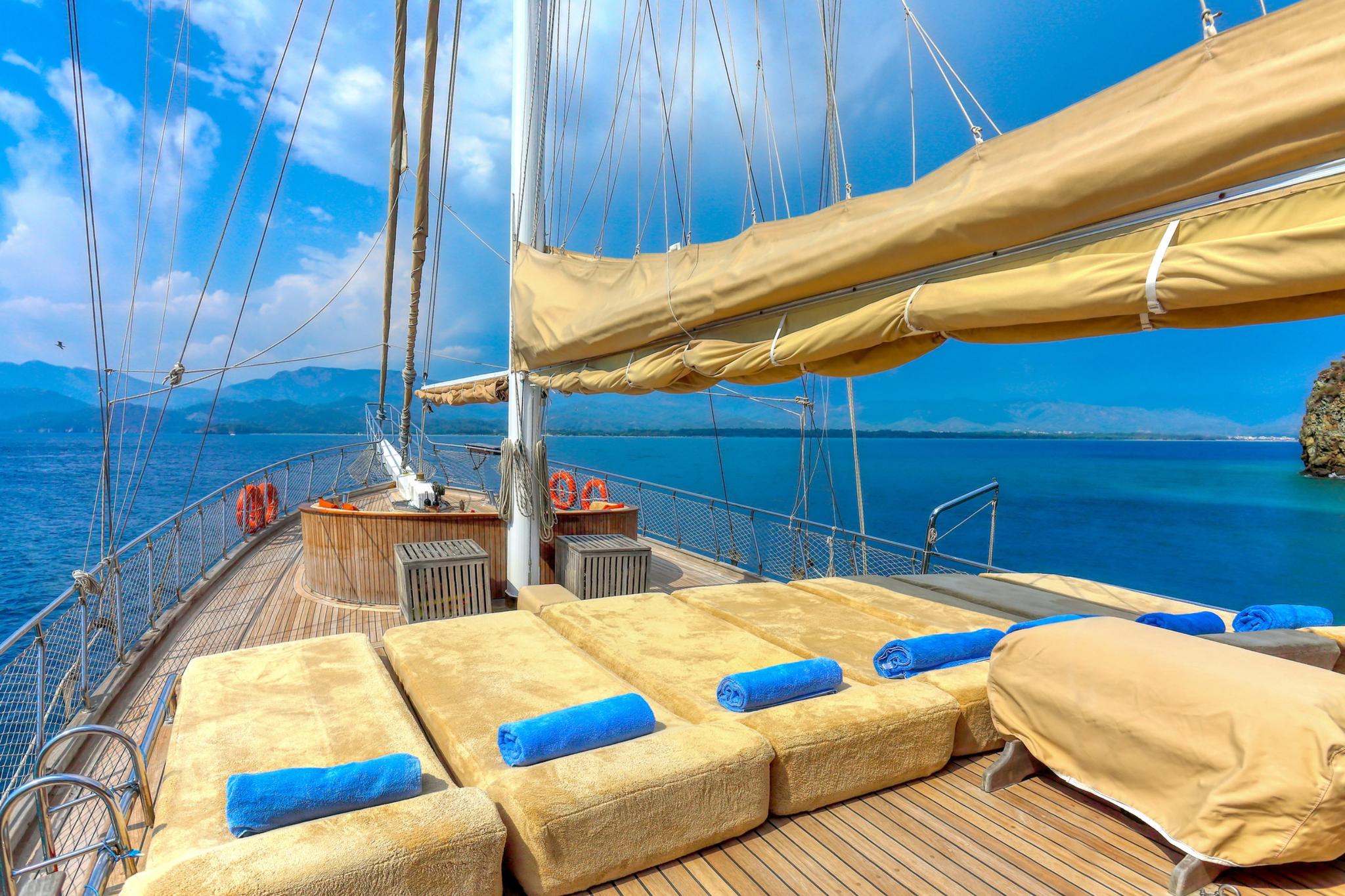 Gulet bow sun deck with beige cushioned loungers, blue towels, and cream sails overhead