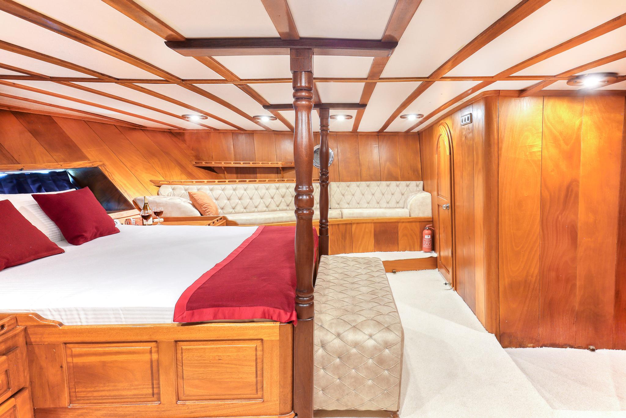 Guest cabin featuring double bed with red runner, cream diamond-quilted banquette seating, and rich teak woodwork