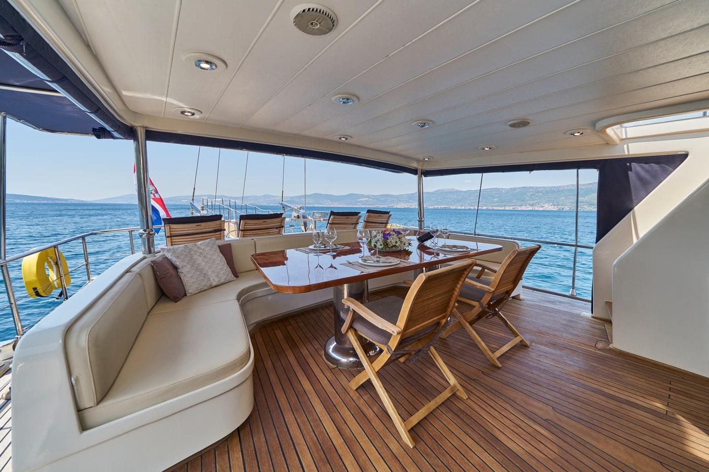 yacht covered aft deck with dining table, curved seating and teak flooring overlooking blue ocean