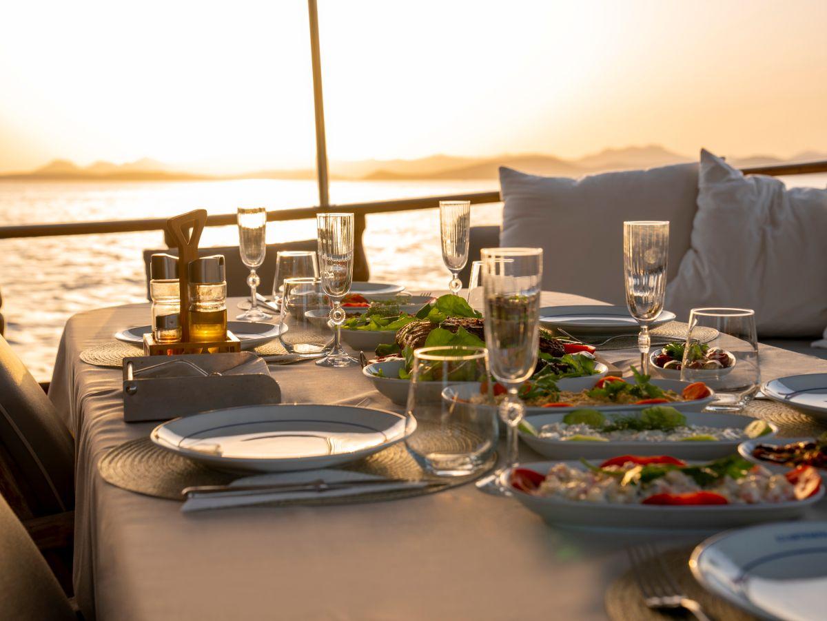 yacht aft deck dining table with prepared meals, wine glasses and cushioned seating at sunset