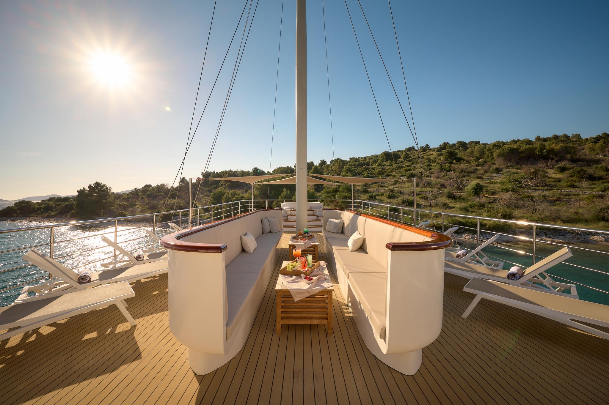 Upper deck seating area with curved white sofas, wooden dining table, and scenic coastal views at sunset