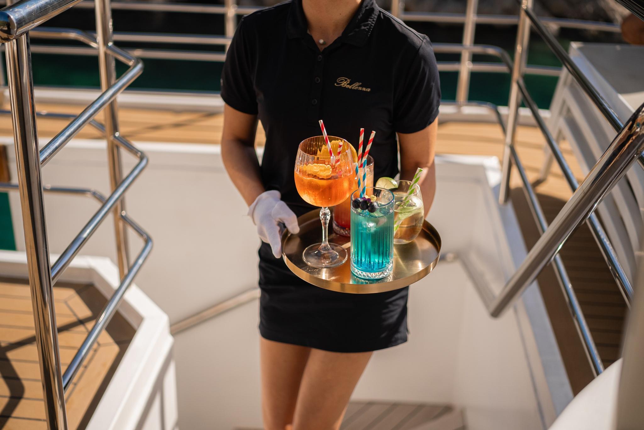 yacht crew member in black uniform serving colorful cocktails on silver tray aboard luxury gulet