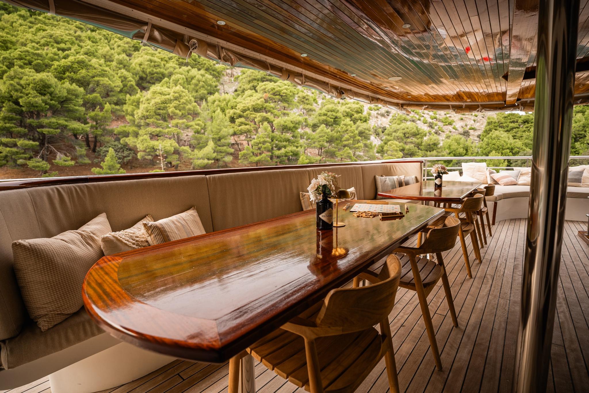 yacht aft deck dining area with wooden table, bench seating and forest coastline views