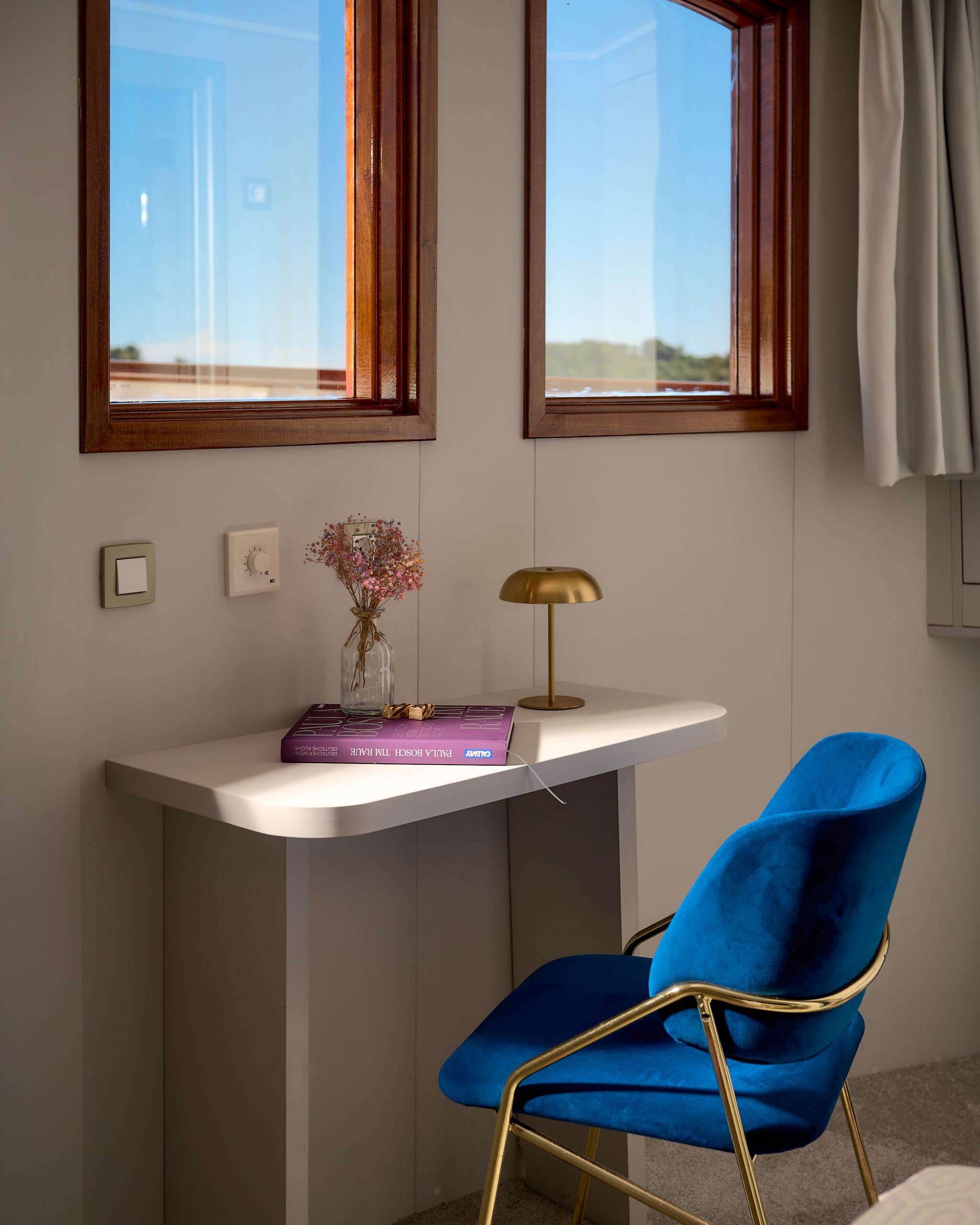 yacht cabin interior featuring white desk, blue velvet chair, two wooden-framed windows