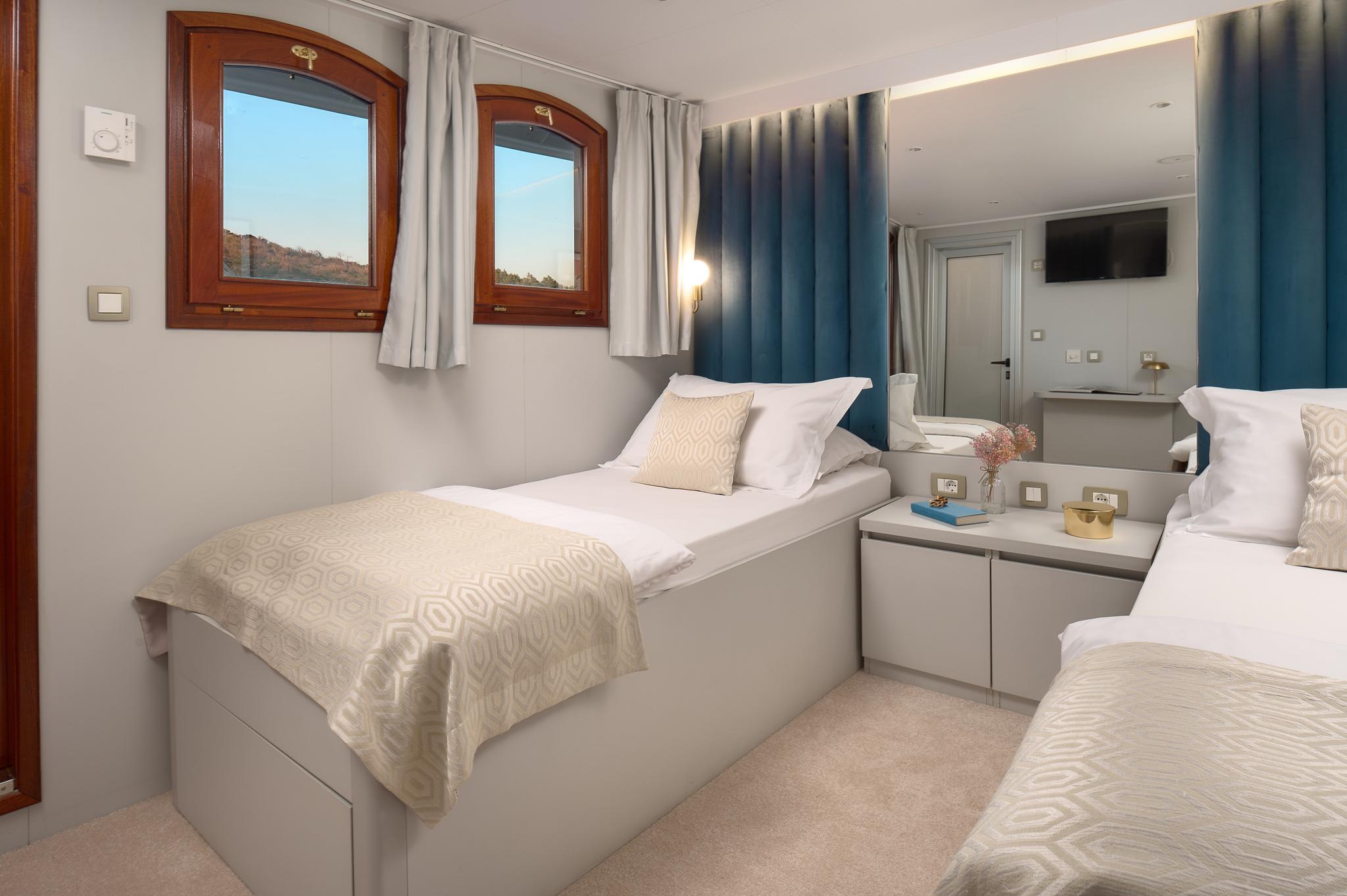 yacht twin cabin with two single beds, blue curtains, and wooden portholes showing coastal view