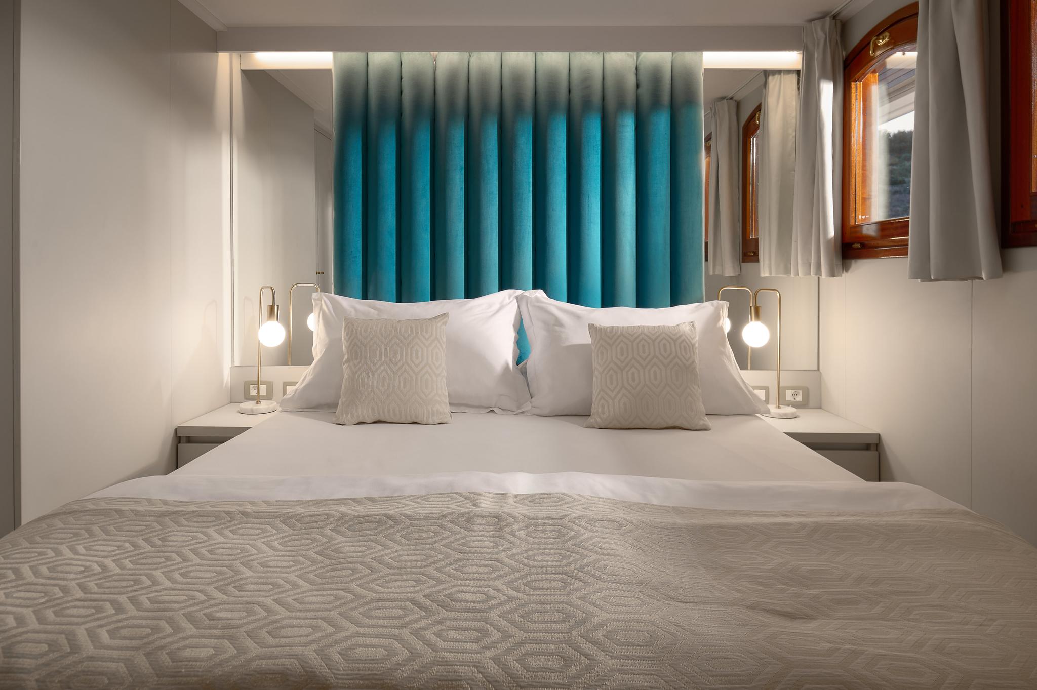 yacht cabin interior with double bed, white linens, turquoise curtains and brass reading lights