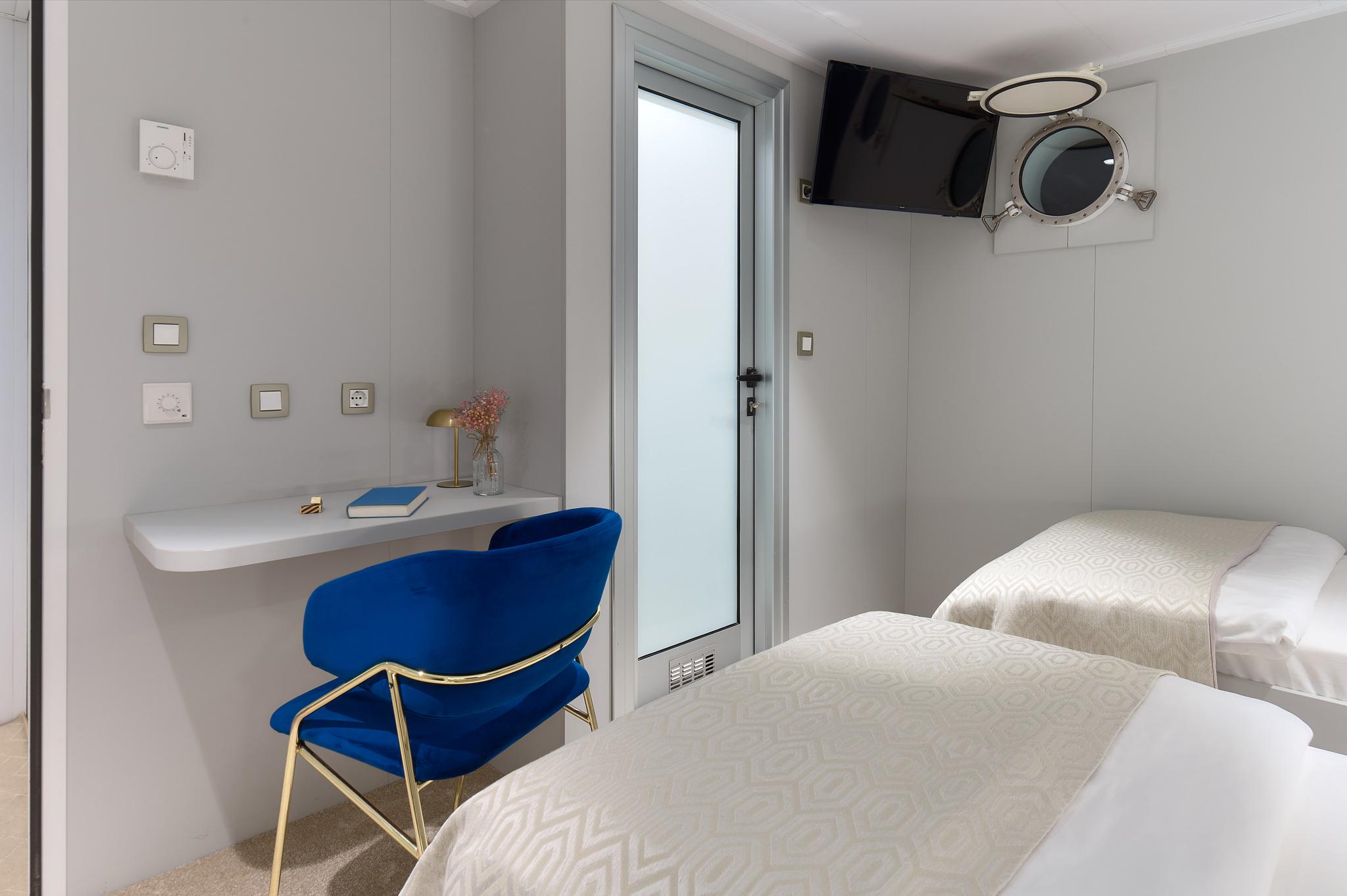 yacht guest cabin with twin beds, blue velvet chair, wall-mounted TV and porthole window
