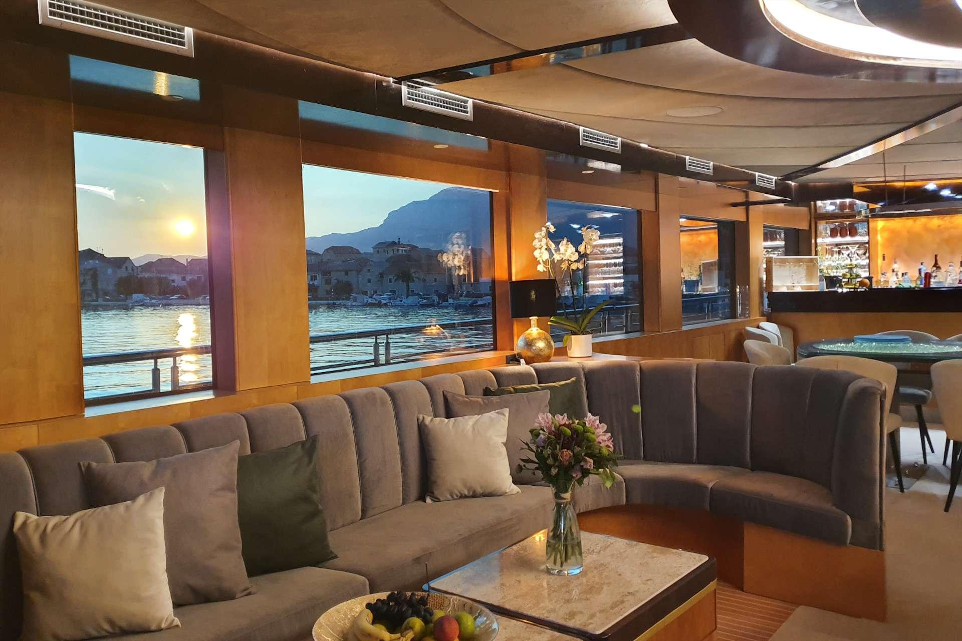 Luxury gulet indoor salon with curved grey velvet sofa, marble coffee table, and built-in bar overlooking a sunset harbour