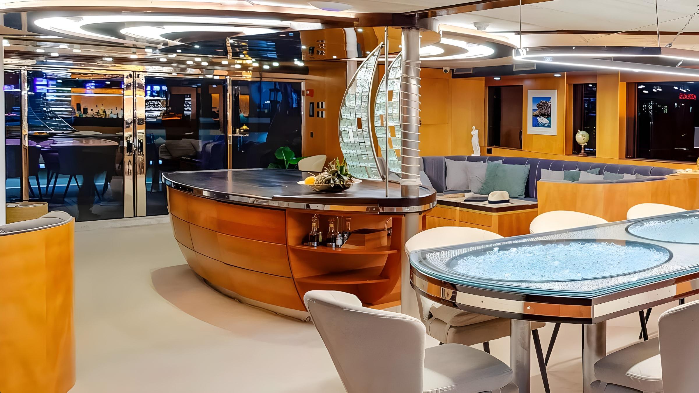 Luxury yacht main salon featuring curved wooden bar unit with glass sail sculpture, illuminated oval dining table with crystal inlay, and wraparound blue cushioned seating