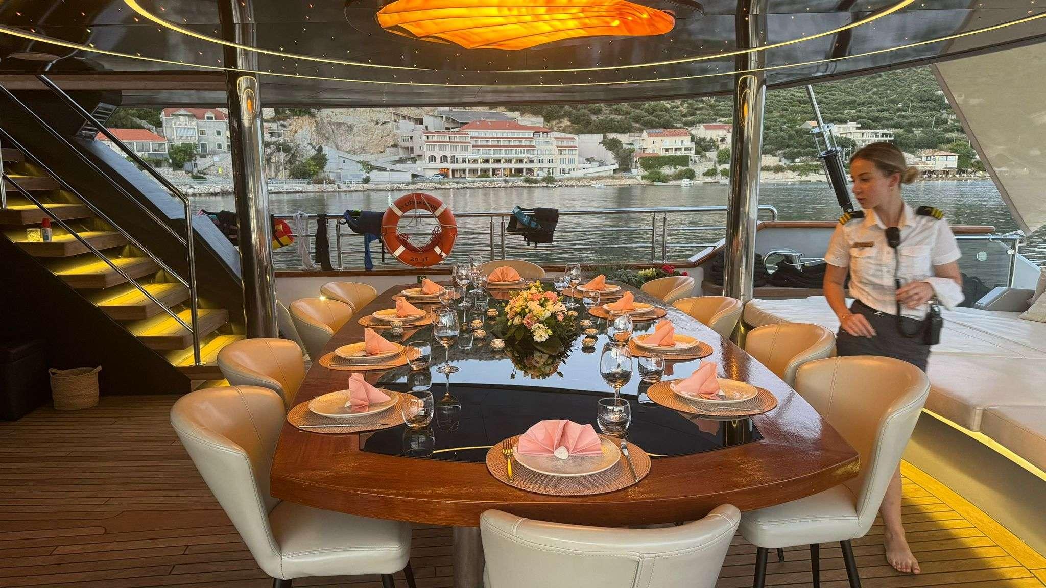 Aft deck dining table formally set for ten with salmon napkins, wine glasses, and floral centrepiece, crew member in uniform attending