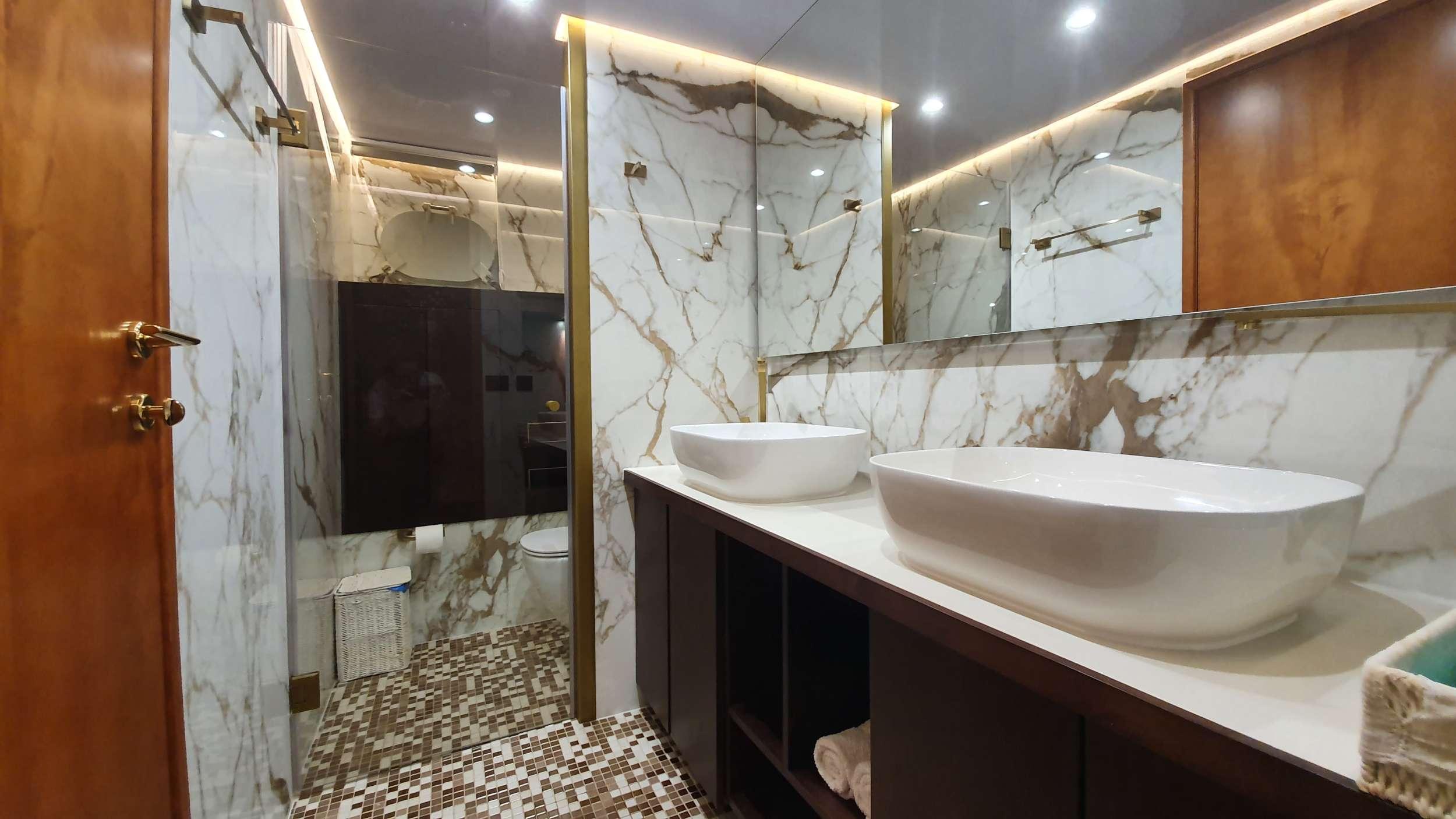 Luxury yacht en-suite bathroom with twin white vessel sinks, Calacatta marble walls, gold fixtures, mosaic tile floor, and glass-enclosed shower area
