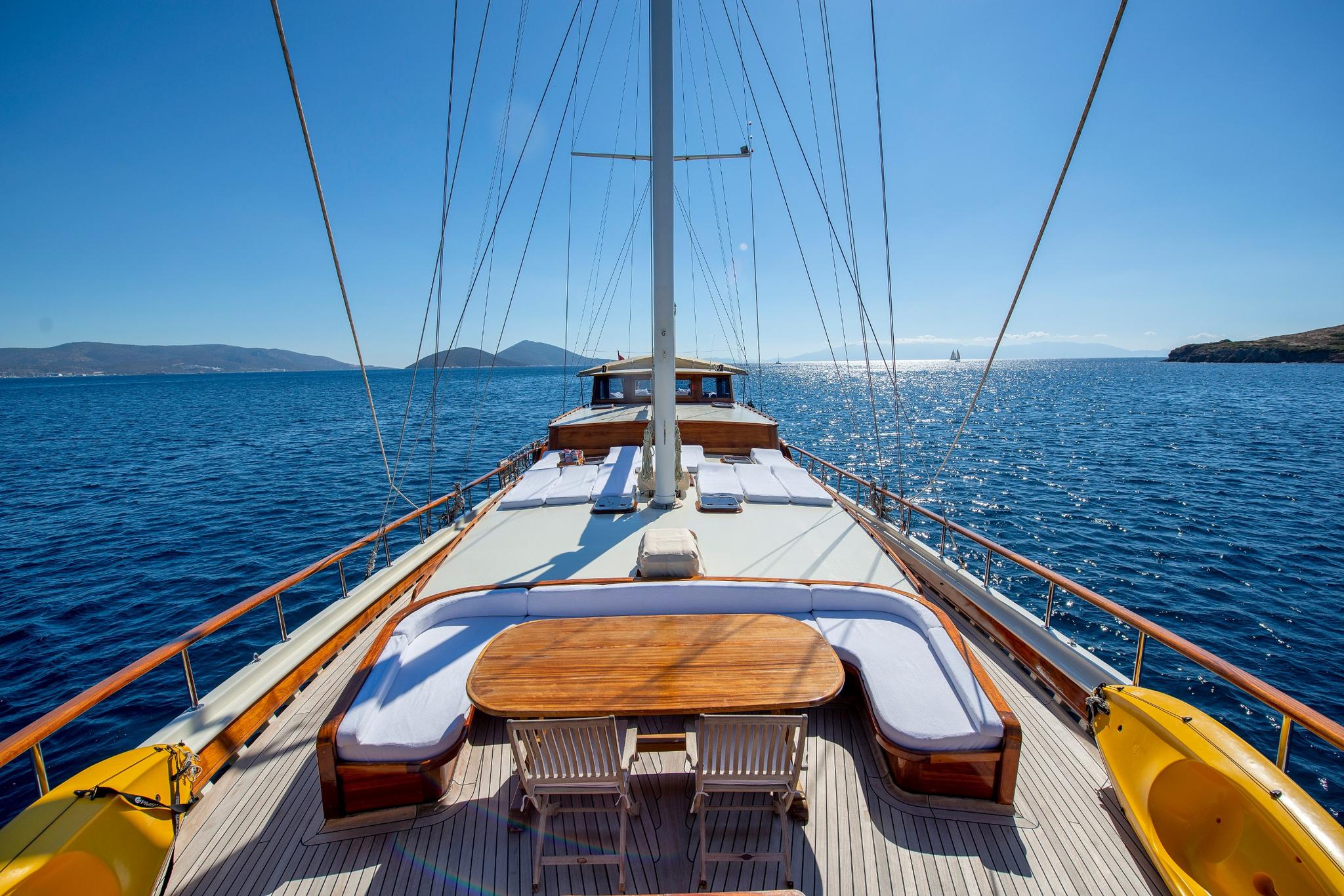 Gulet bow deck with curved cushioned seating around oval teak table, white mast and rigging