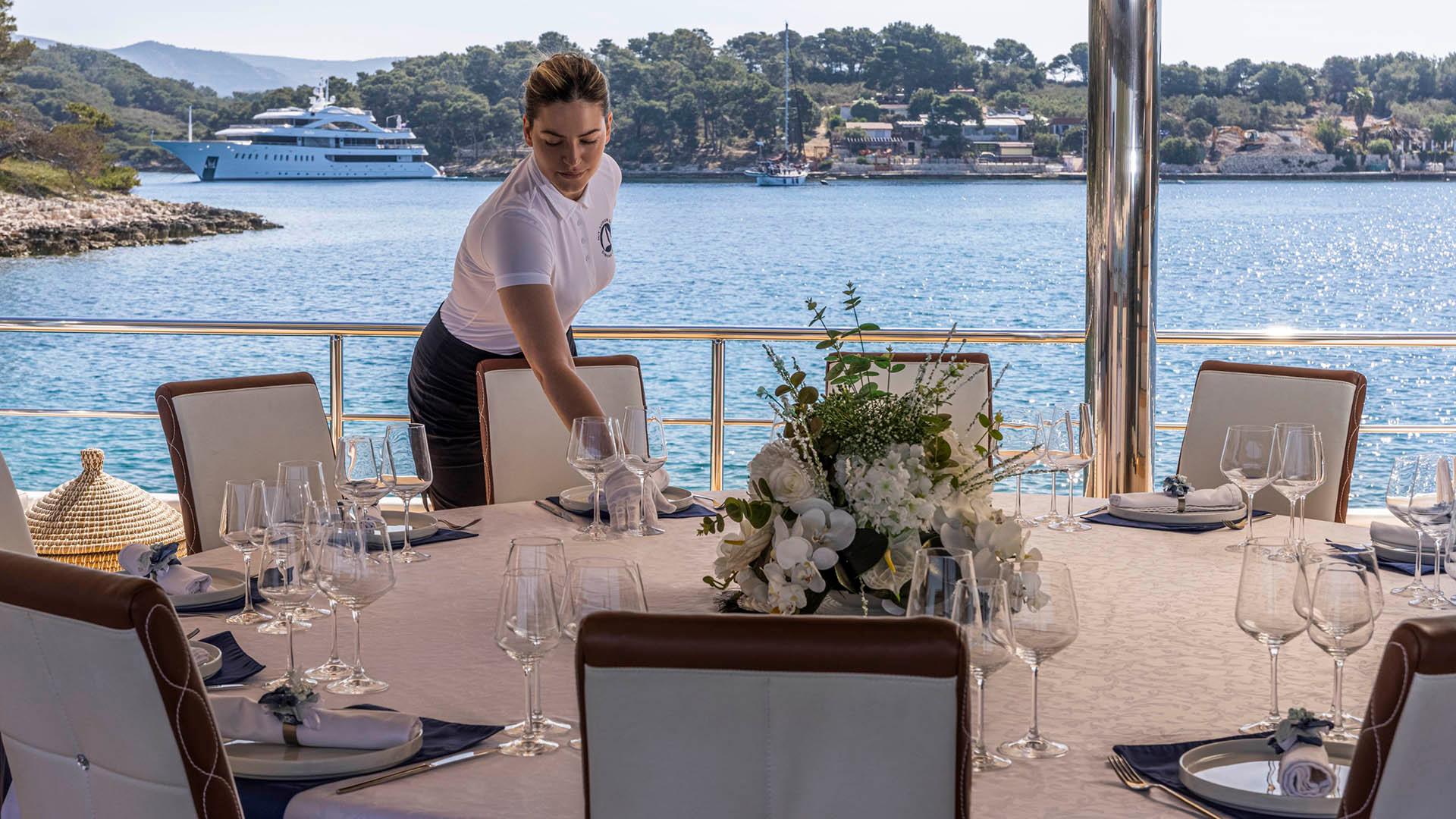 crew member arranging glassware on formal dining table with floral centerpiece on yacht aft deck