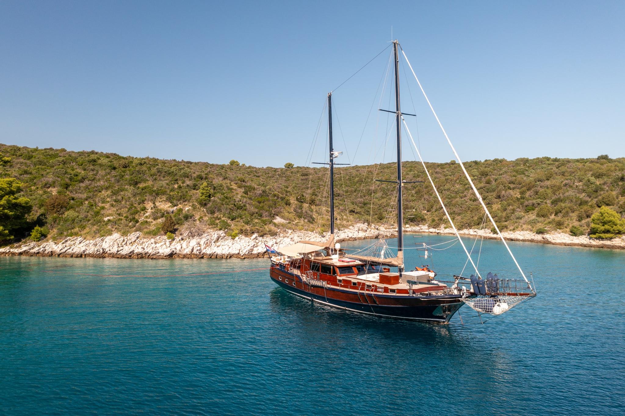 wooden gulet yacht with two masts anchored in crystal clear turquoise bay surrounded by rocky coastline