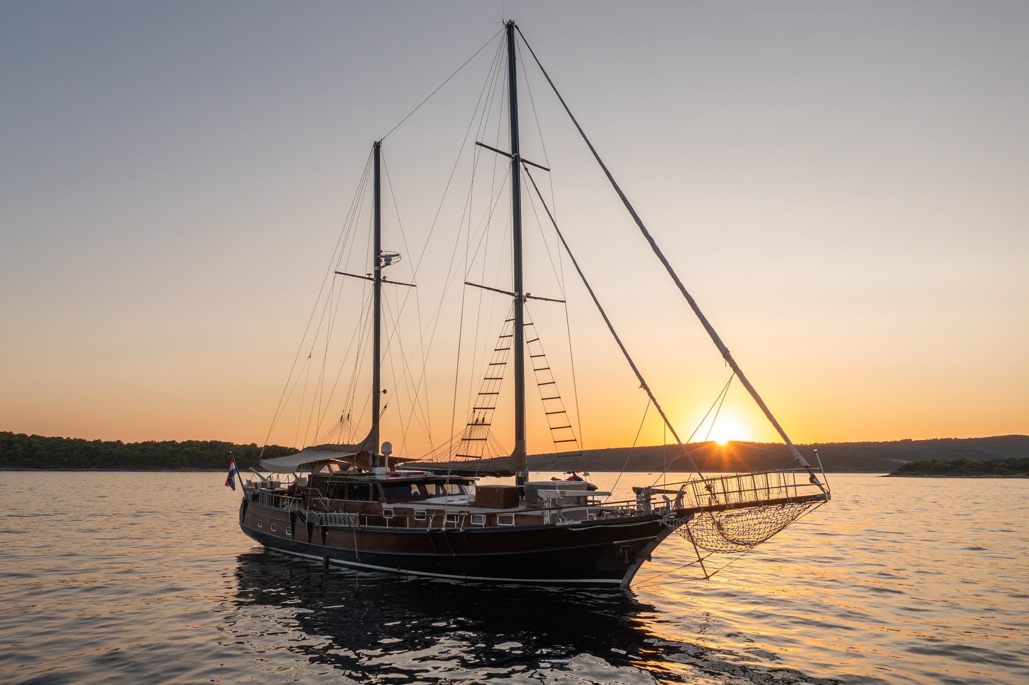 wooden gulet yacht with two masts anchored at golden sunset with forested coastline