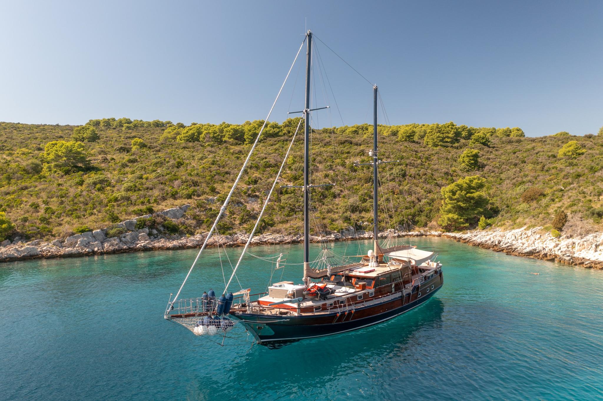 traditional wooden gulet yacht with two masts anchored in crystal clear turquoise bay surrounded by forested coastline