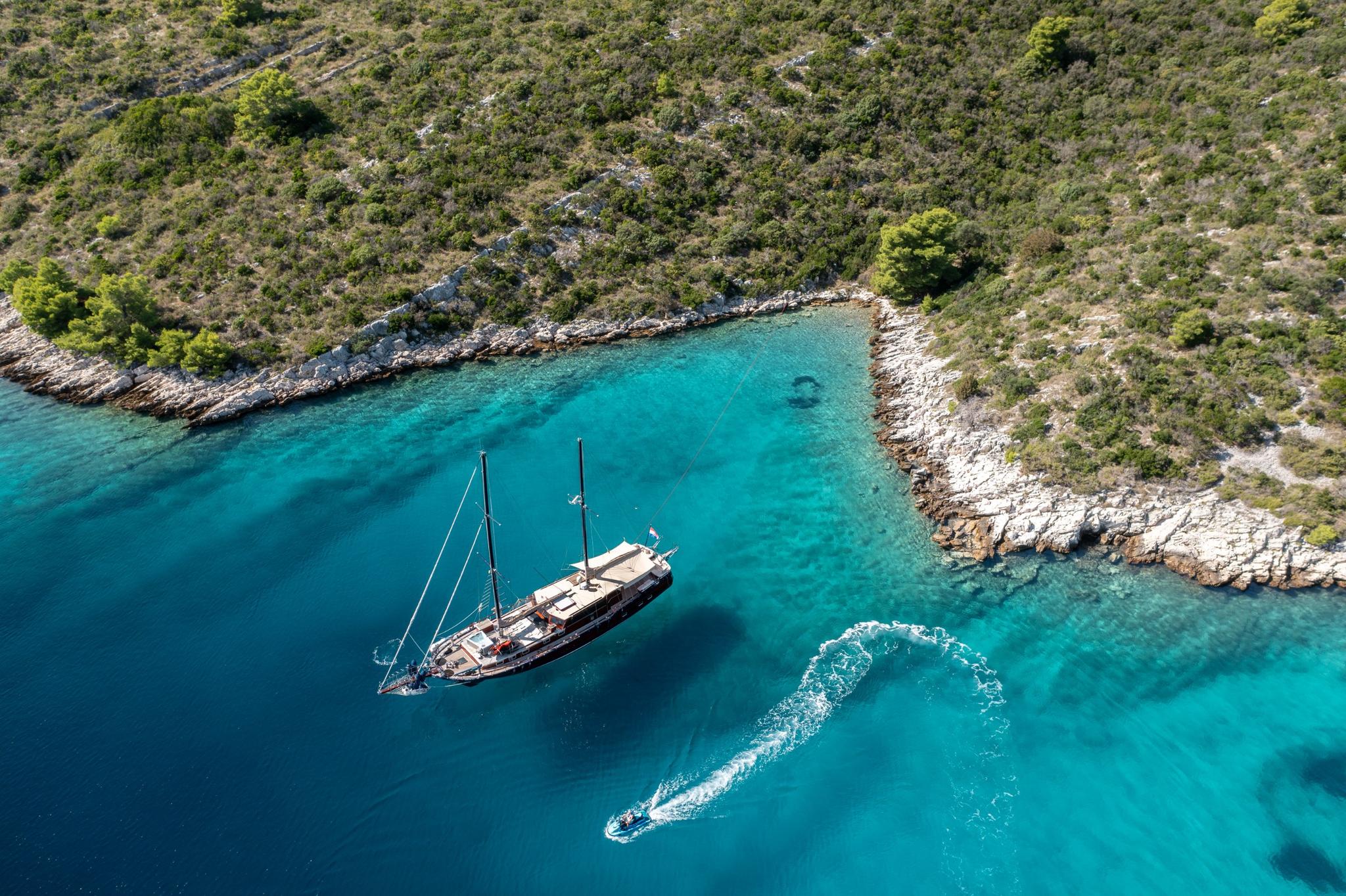 aerial view of traditional wooden gulet yacht anchored in crystal clear turquoise bay with tender boat creating wake