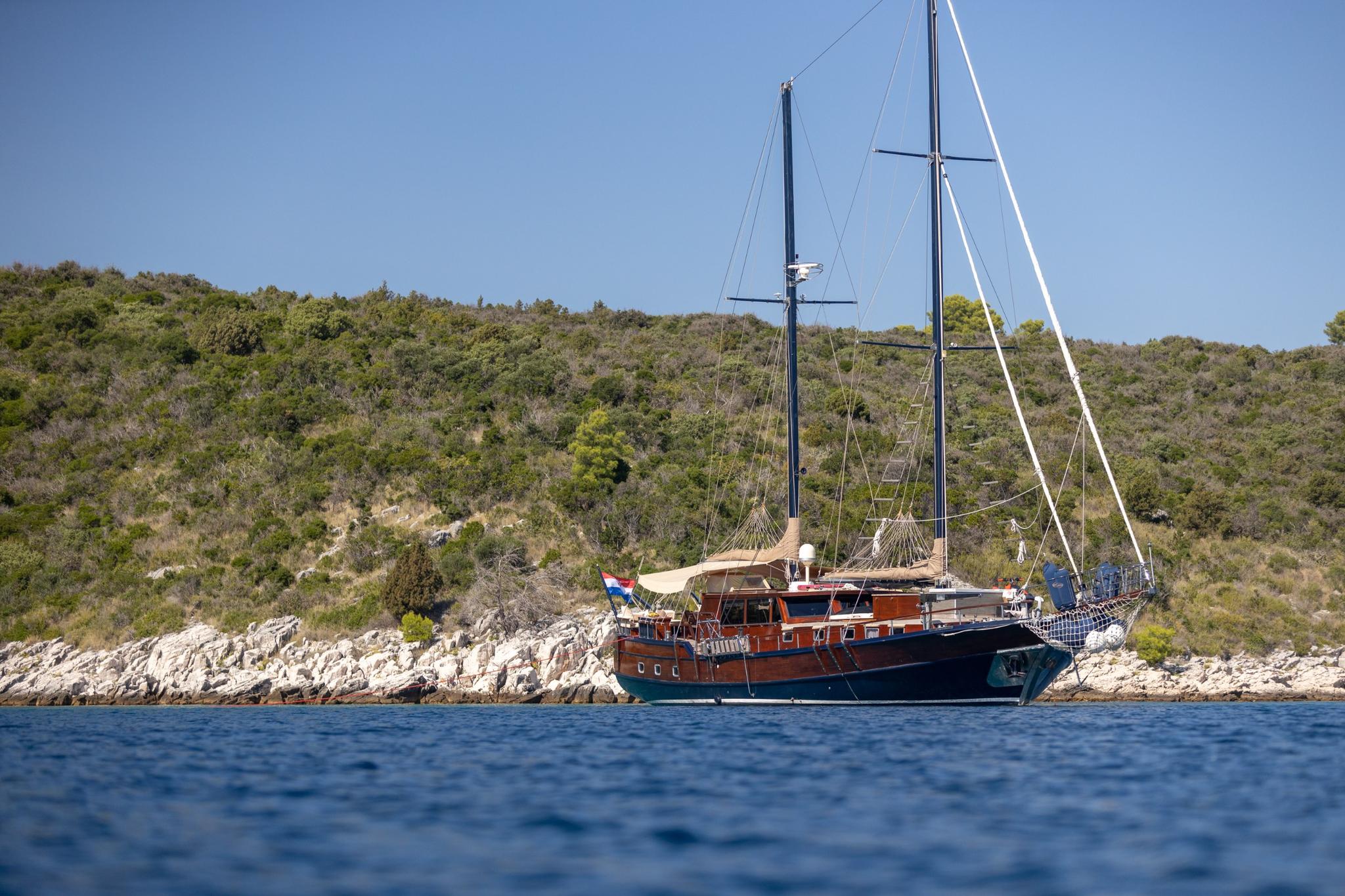 wooden gulet yacht with two masts anchored in blue waters near Mediterranean rocky coast
