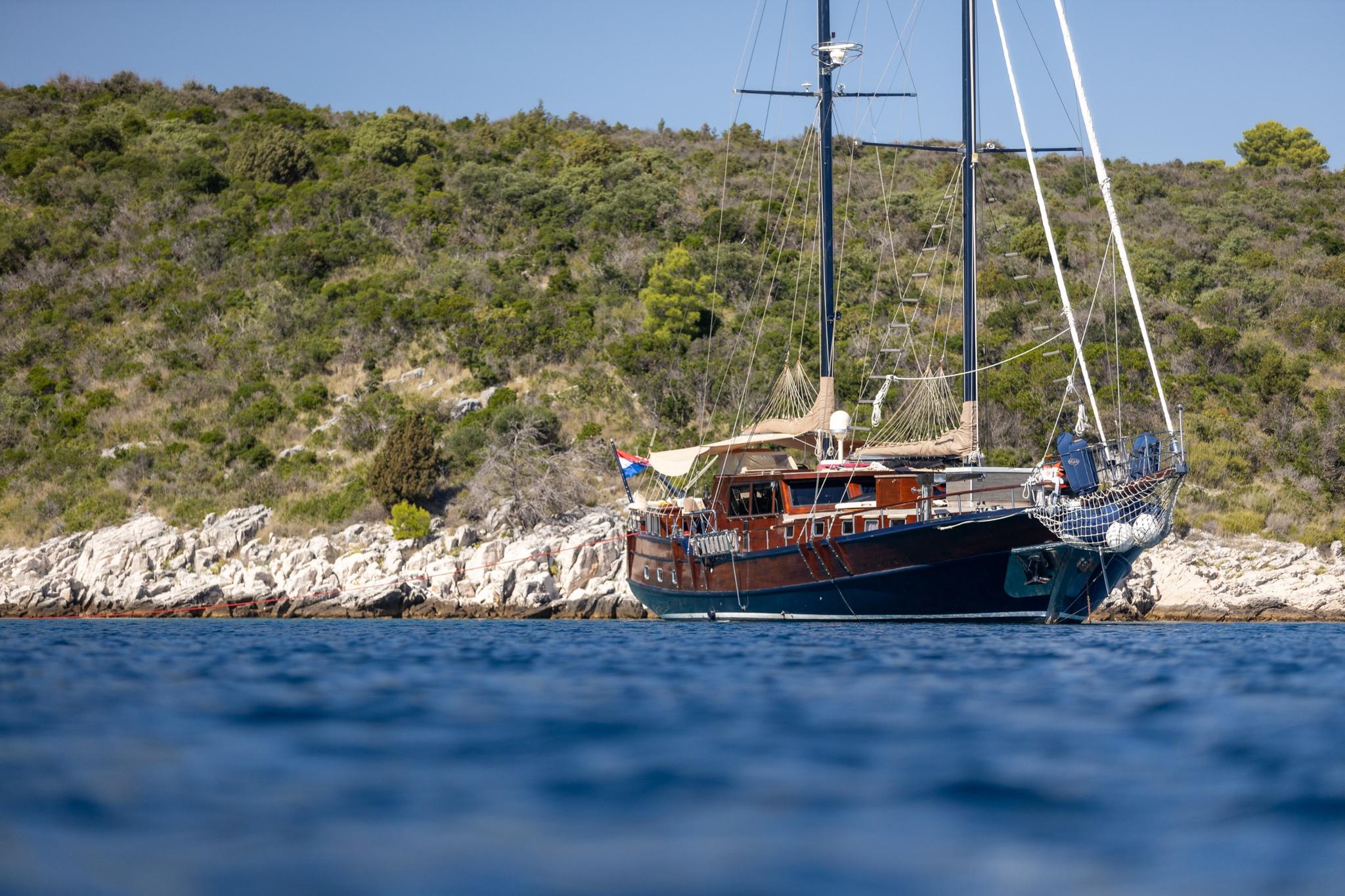 wooden gulet yacht with two masts anchored in blue waters near Mediterranean rocky coastline