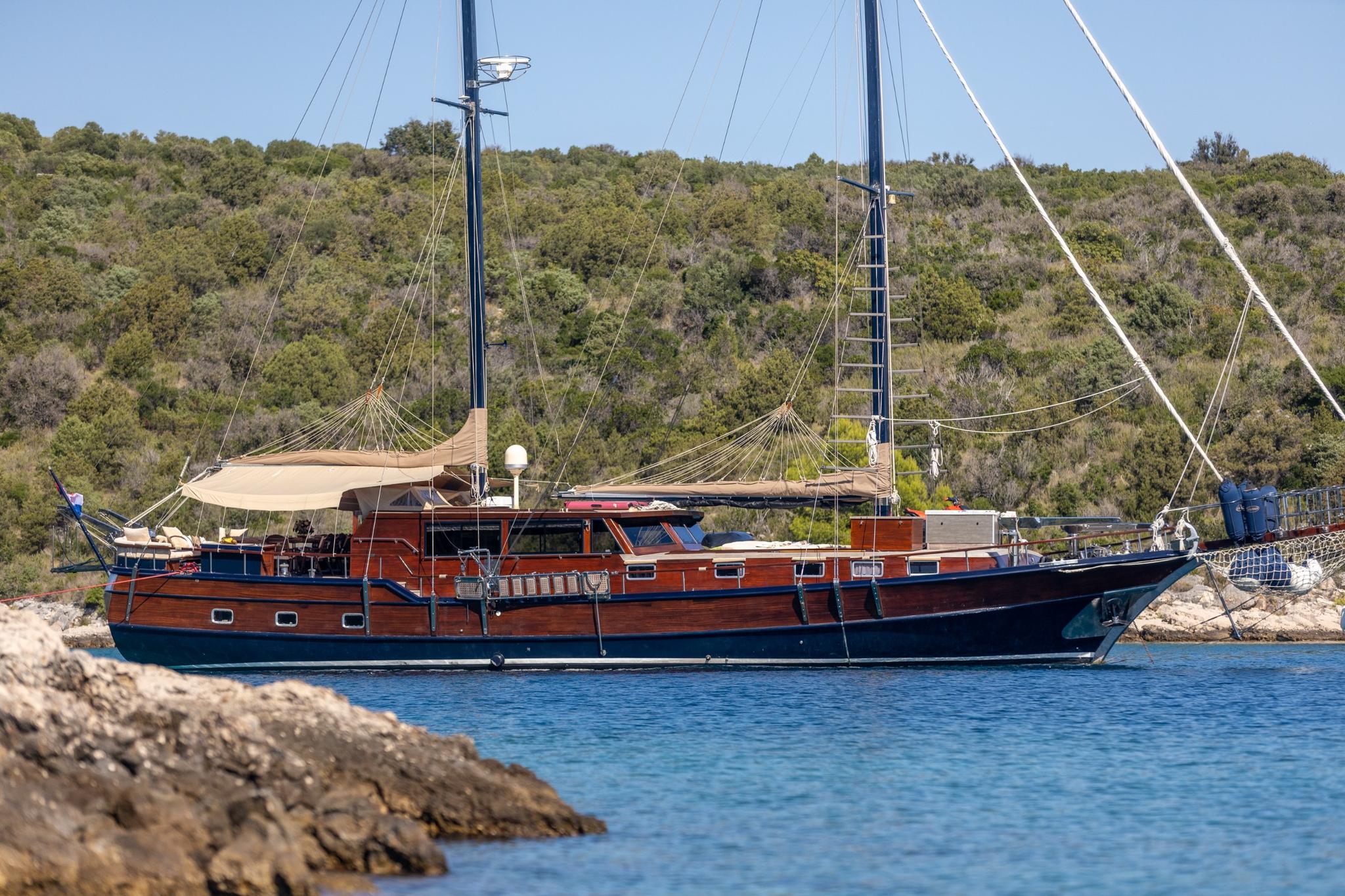wooden gulet yacht with dark blue hull anchored in turquoise Mediterranean waters near rocky coastline