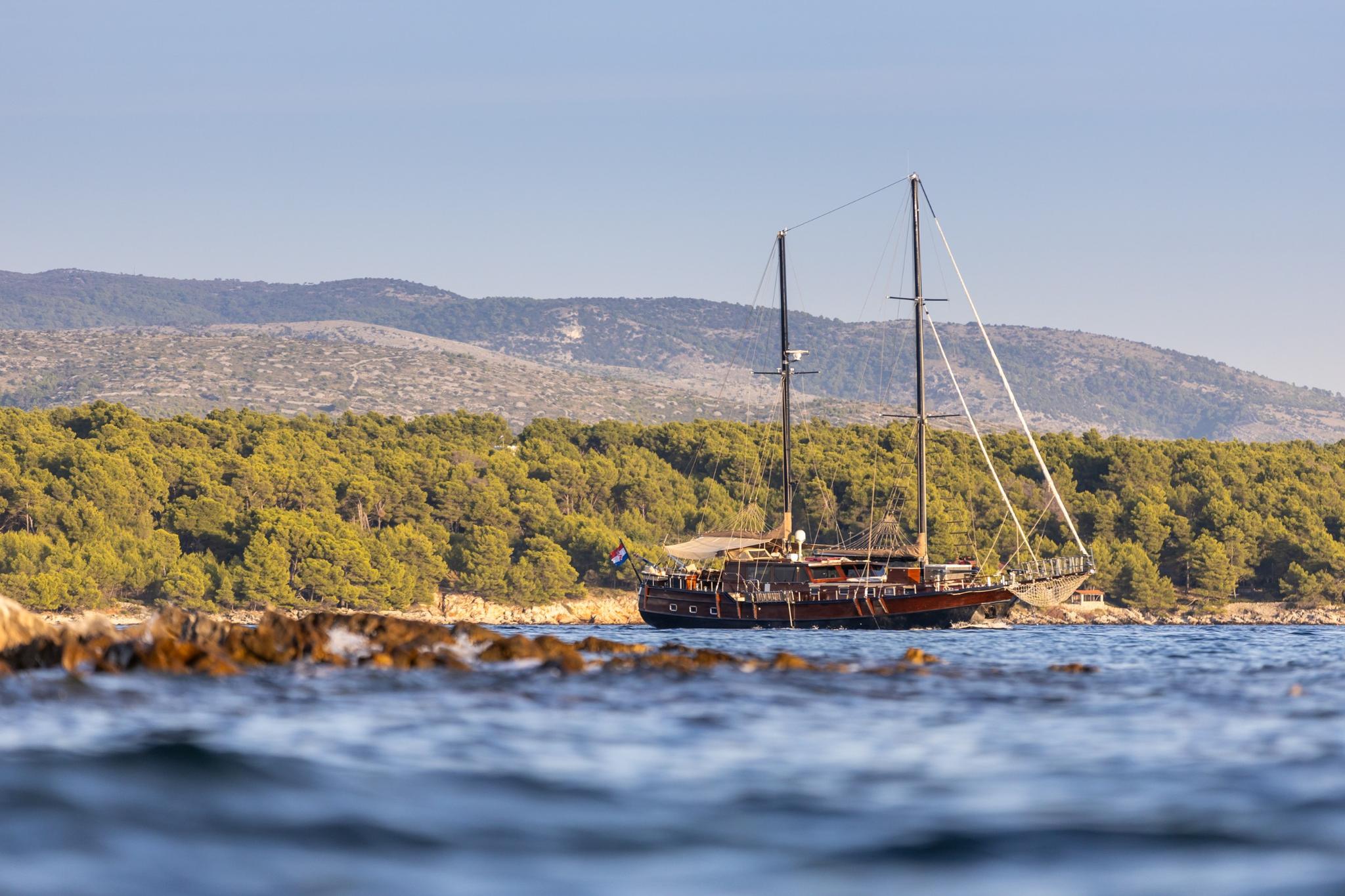 wooden gulet with two masts anchored near pine-forested Mediterranean coastline with mountains