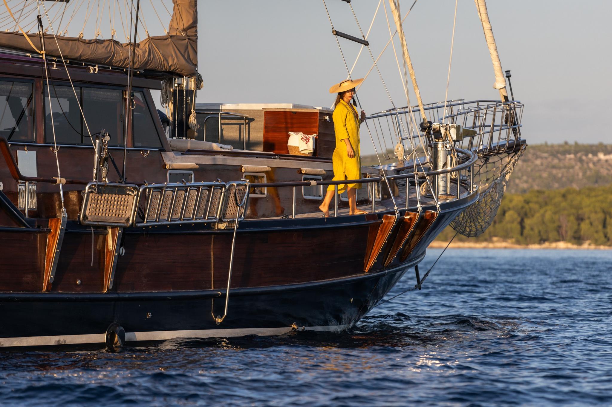 wooden gulet bow with guest in yellow dress standing on deck, coastline background