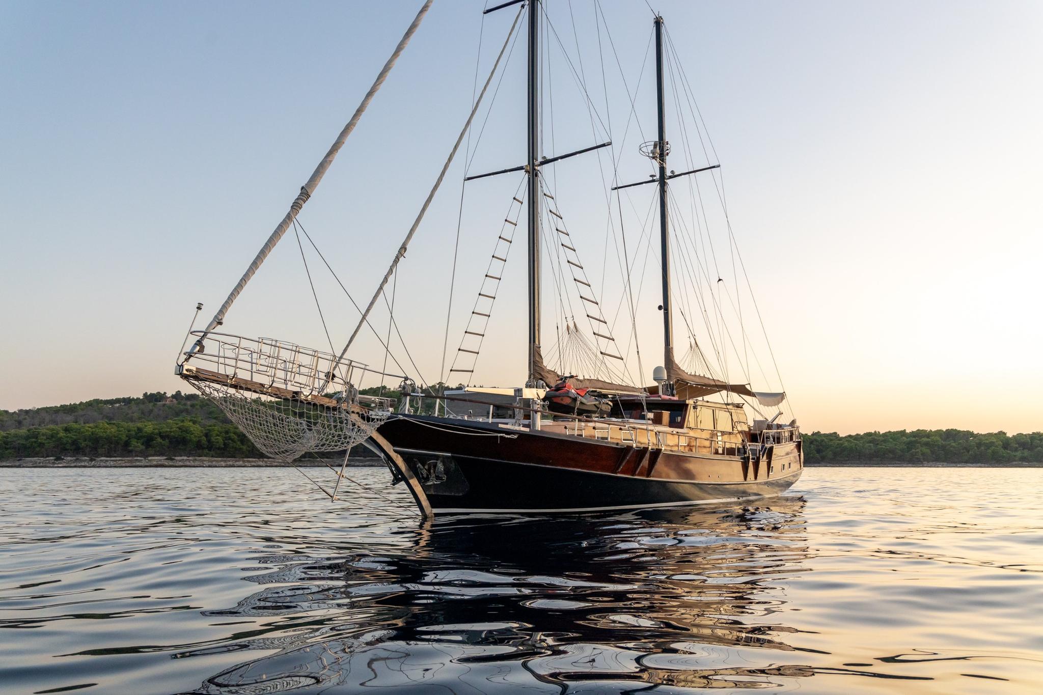 wooden gulet yacht with two masts anchored at sunset with wooded coastline background