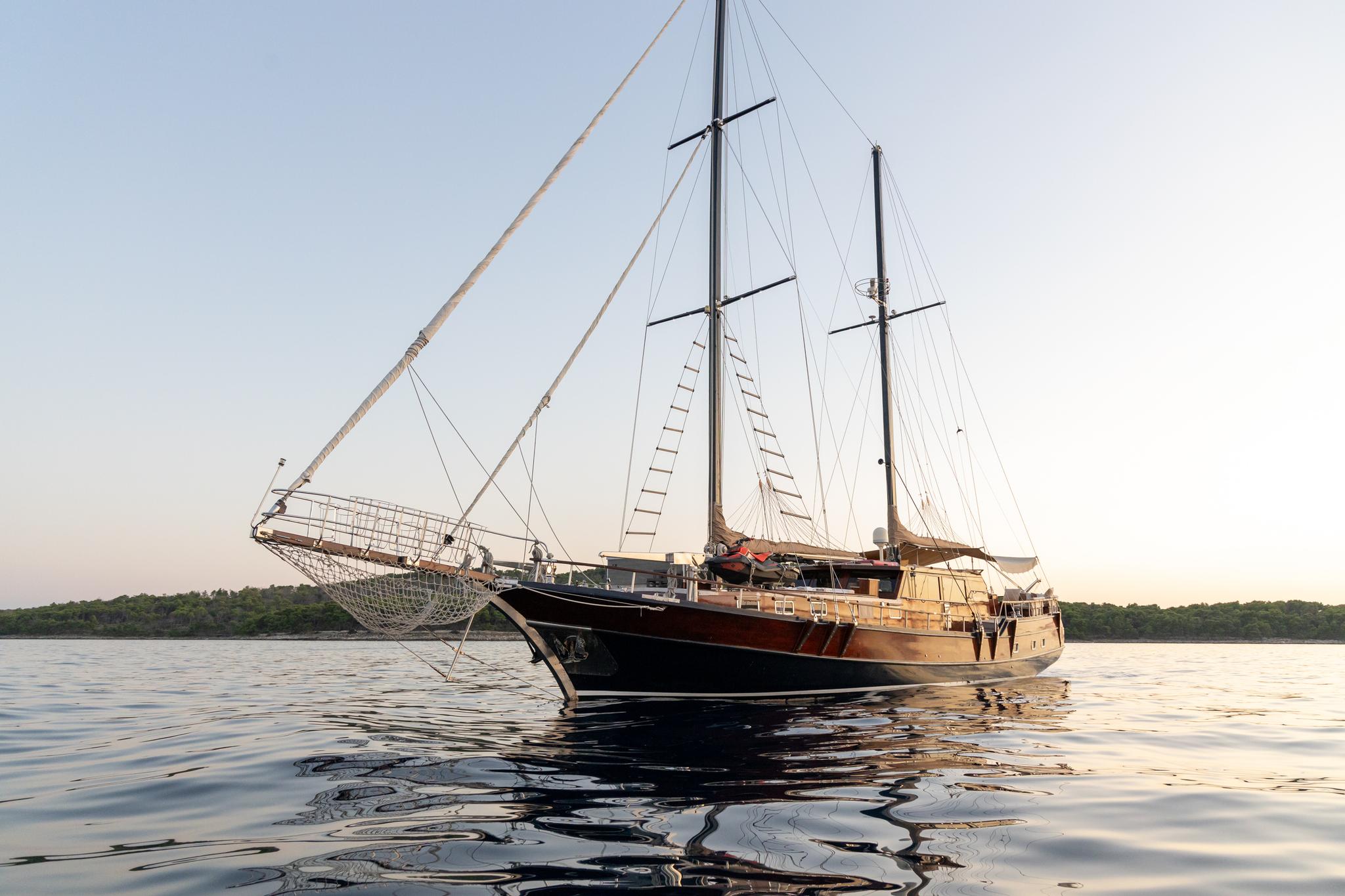 Traditional wooden gulet with two masts and white sails anchored on calm water at sunset