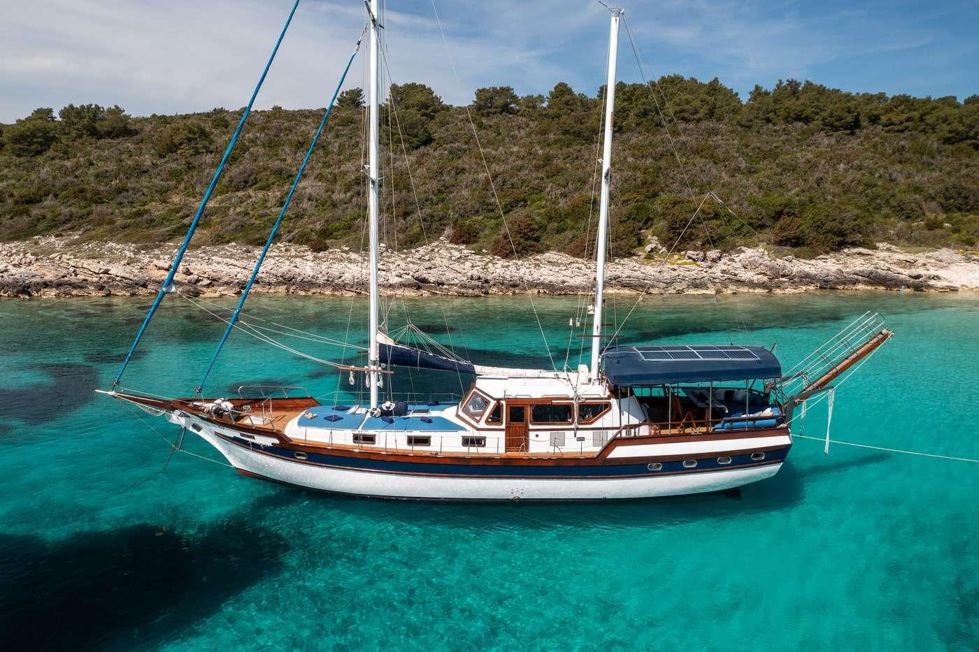 white and wooden gulet with two masts anchored in crystal clear turquoise waters near rocky coastline