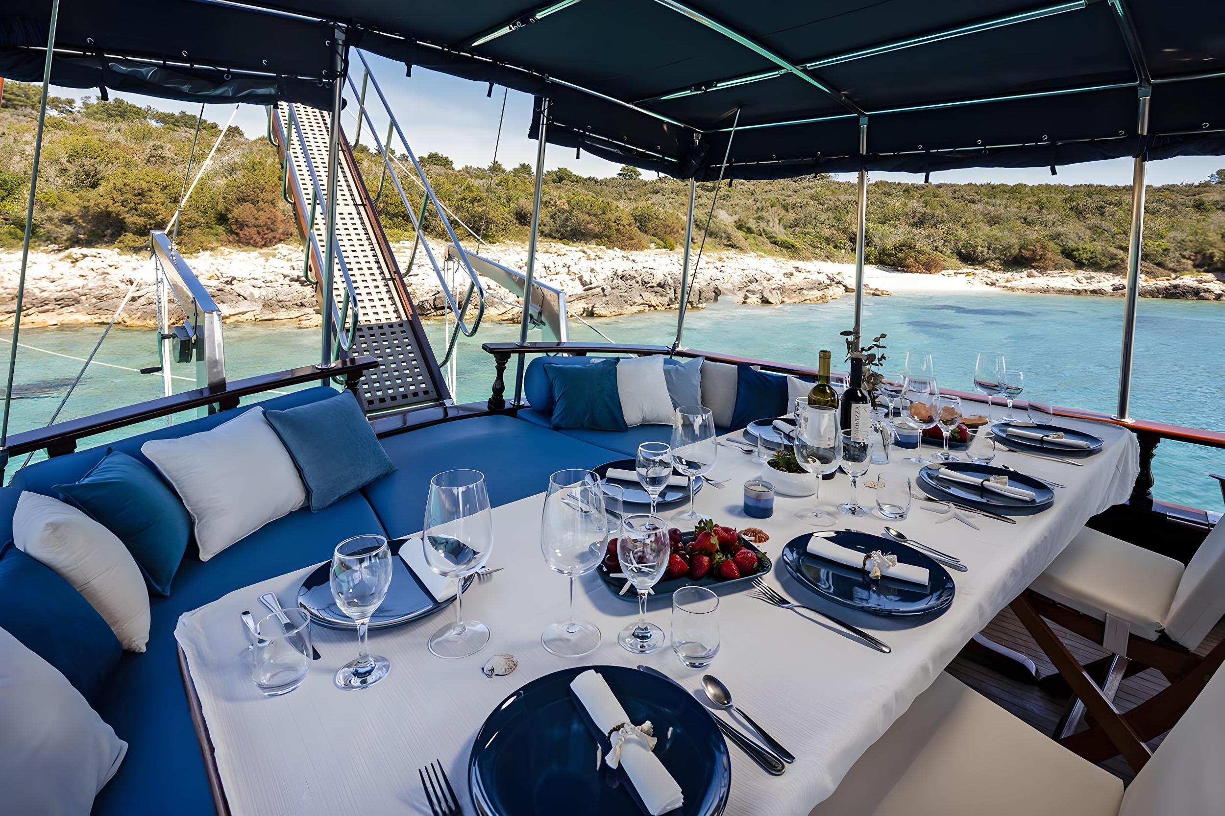 Gulet aft deck dining table set with navy plates, wine bottles, strawberries and crystal glassware anchored in turquoise cove