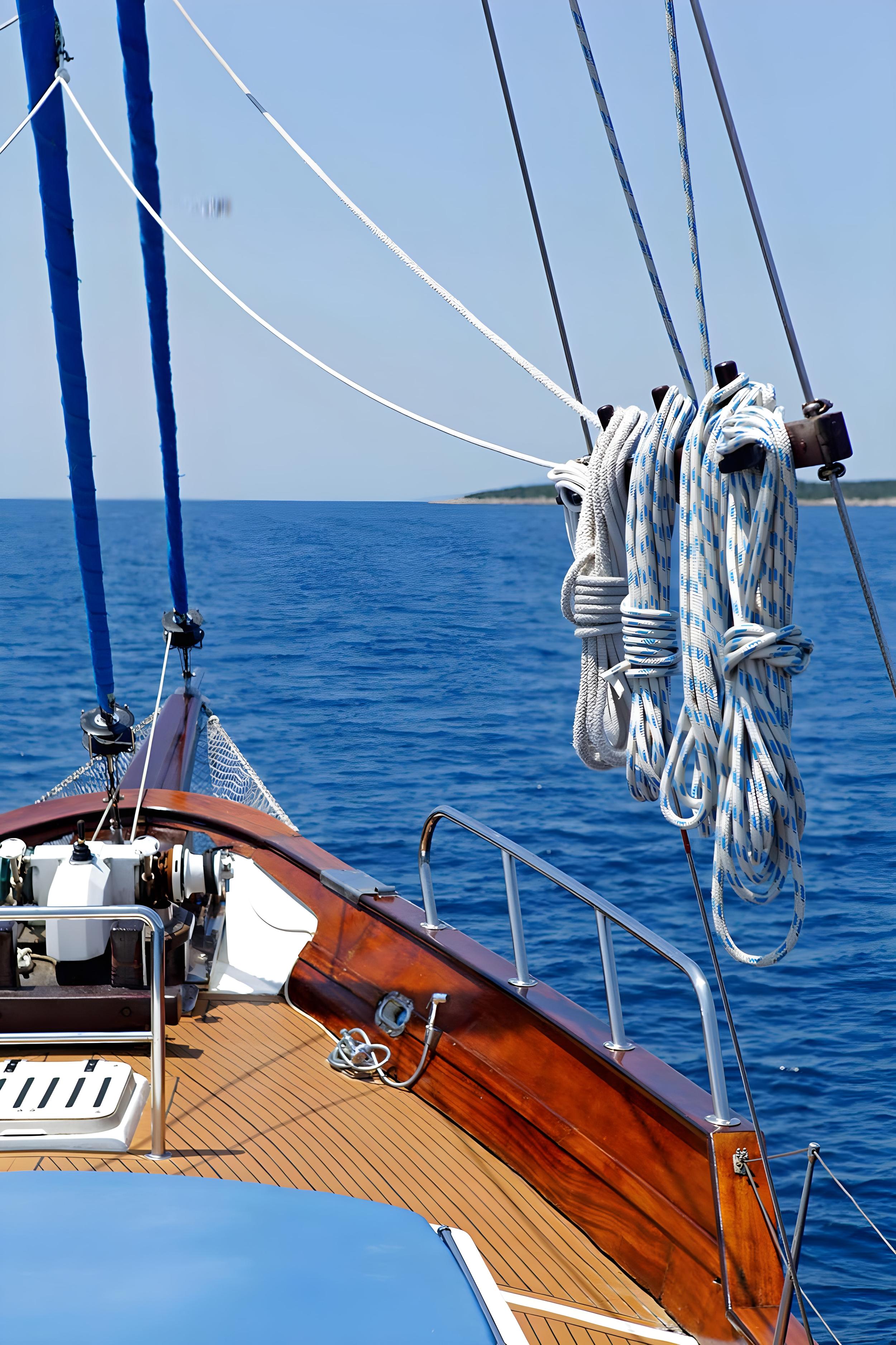 Gulet bow with varnished wooden bowsprit, coiled blue-white ropes on rigging, and teak deck overlooking calm blue sea