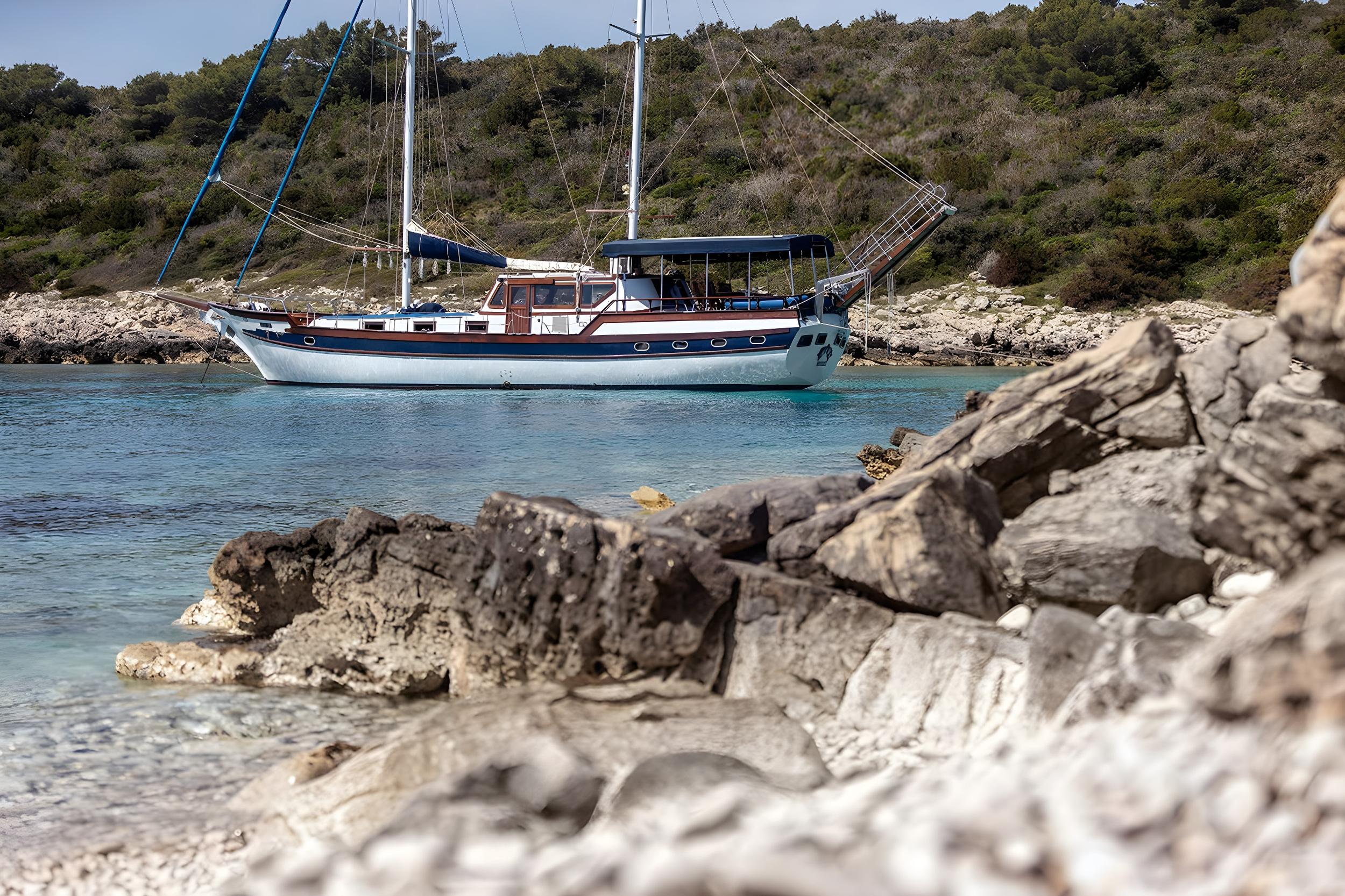 Traditional wooden gulet with blue hull anchored in clear turquoise waters beside rocky Mediterranean coastline