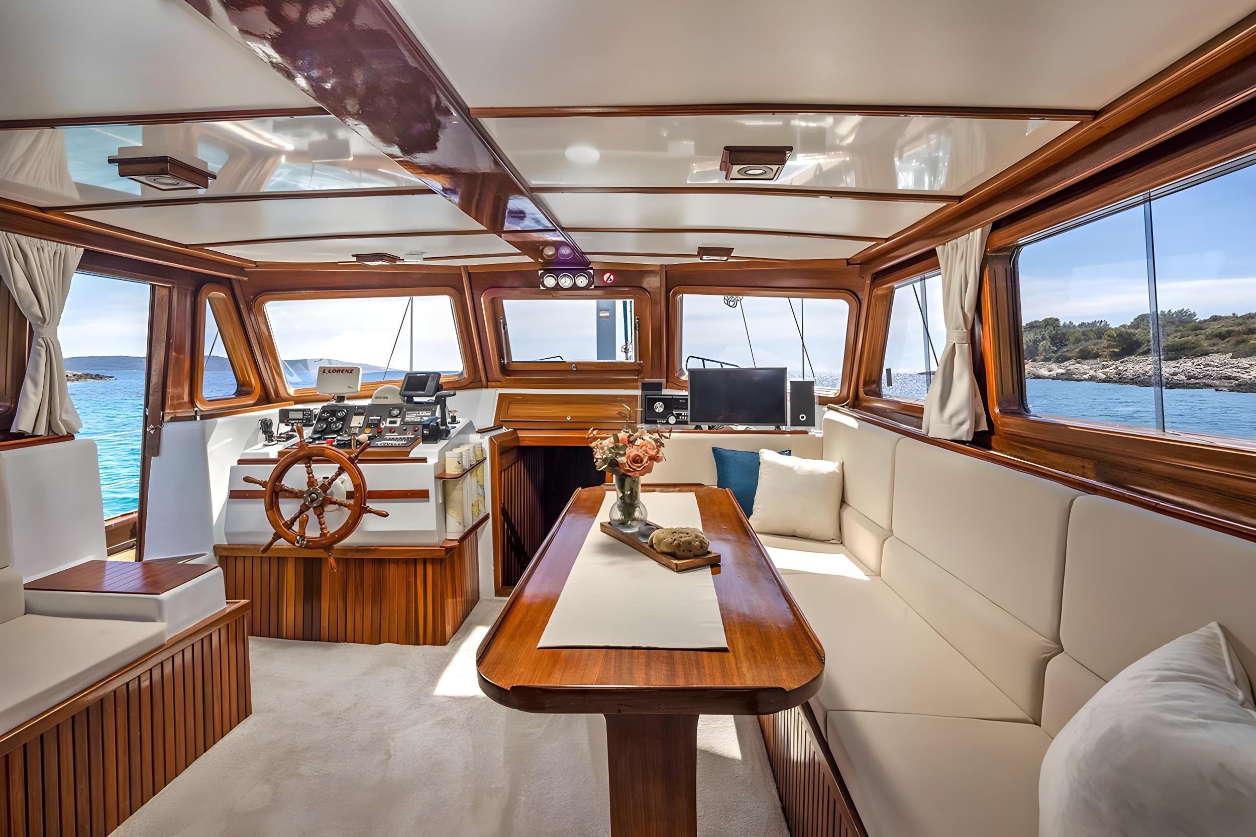 Gulet interior saloon featuring wooden helm wheel, navigation instruments, cream upholstered seating, and polished teak dining table with flower arrangement