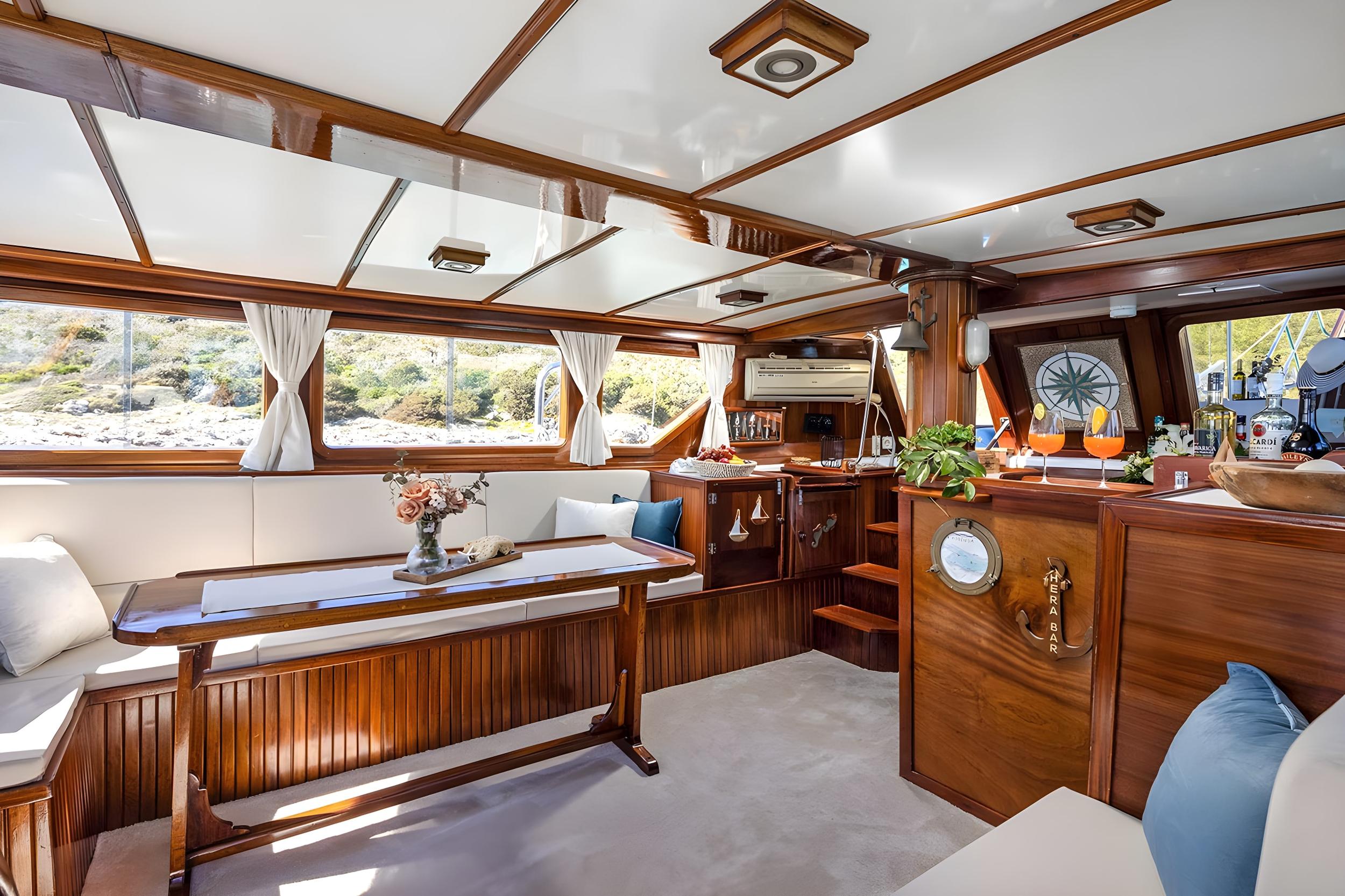Gulet interior saloon featuring polished teak woodwork, built-in bar labelled Hera Bar, L-shaped seating, and dining table with floral arrangement